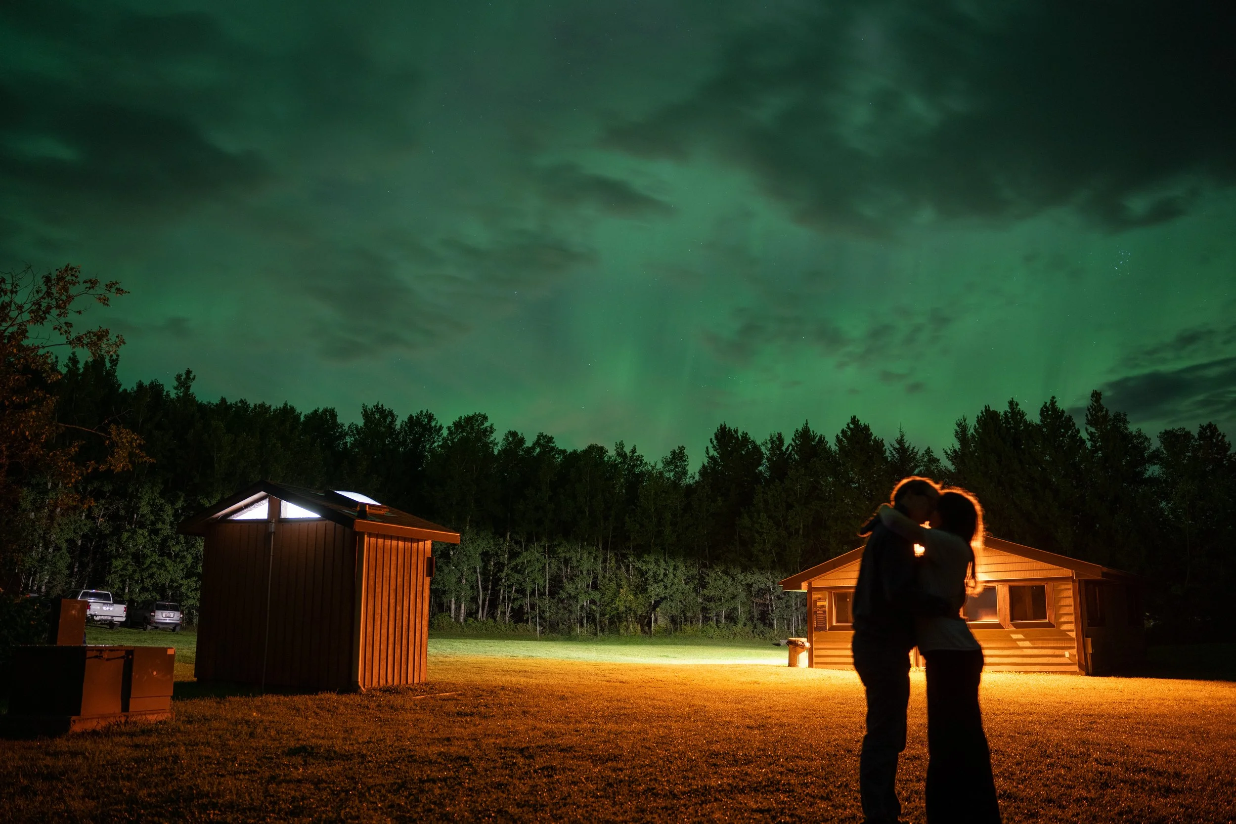 People embracing and kissing outdoors at night with the Northern Lights in the sky.