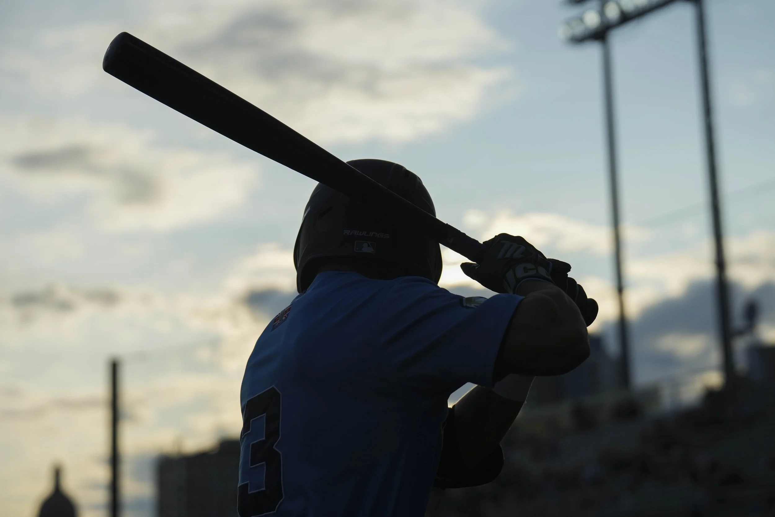 A person holding a baseball bat, dressed in a blue shirt with a name and number on the back, in silhouette against a cloudy sky, with stadium lights in the background.