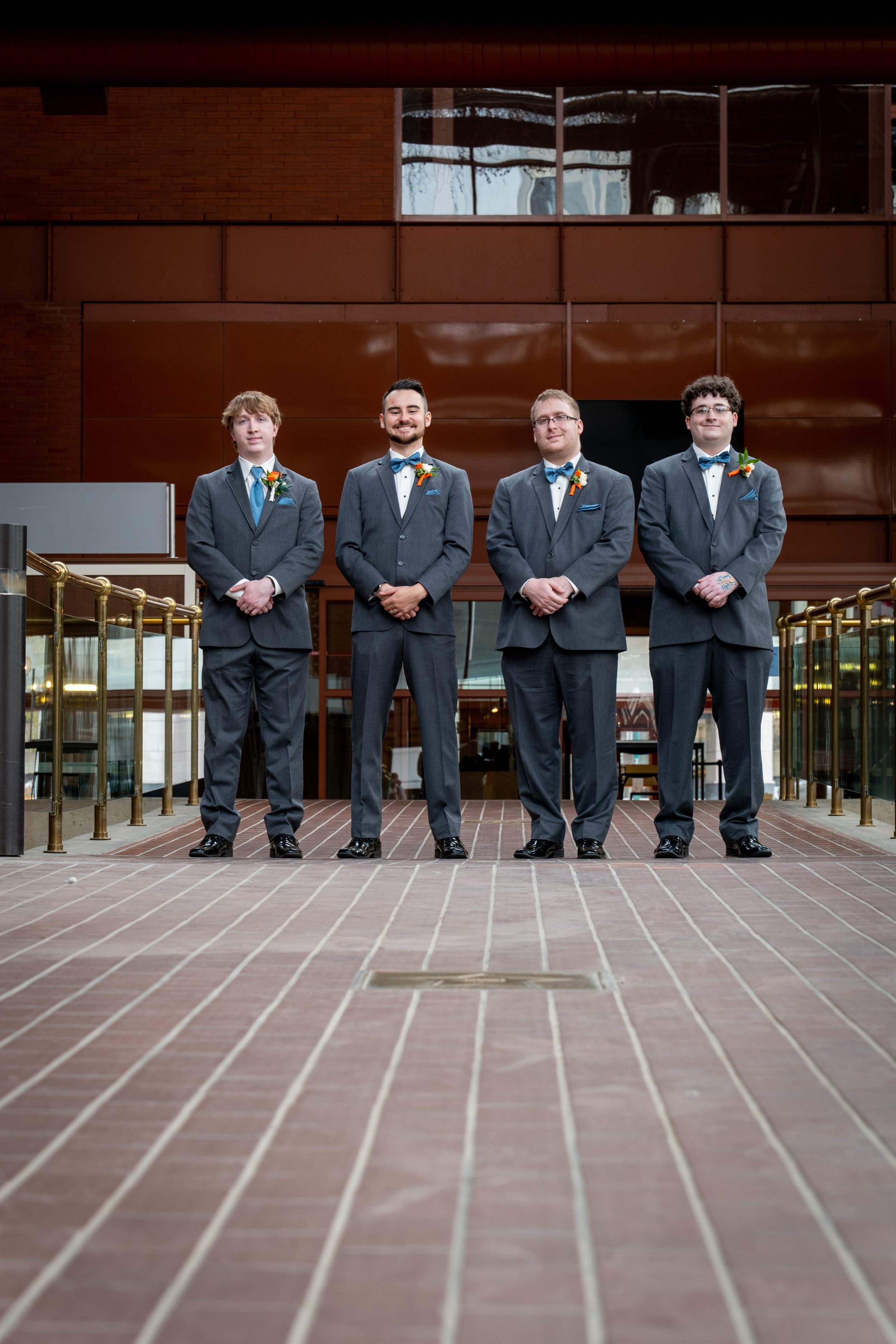 Four men in matching gray suits and blue bow ties standing in a row inside a building with brown walls and glass windows behind them.