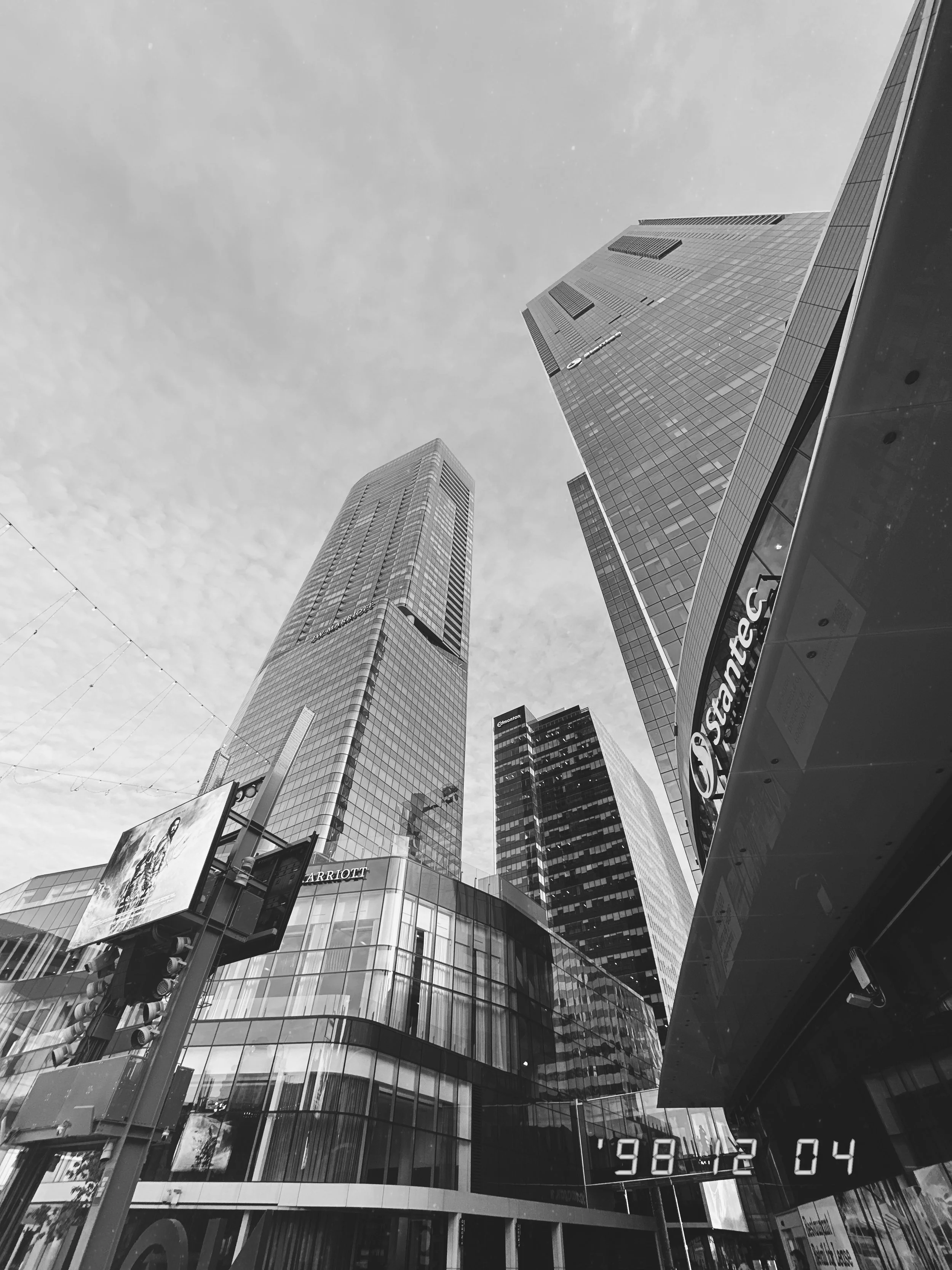 Black and white photograph of tall modern skyscrapers with glass facades in an urban setting. The perspective is from below, looking up at the buildings, with the sky and some clouds visible.