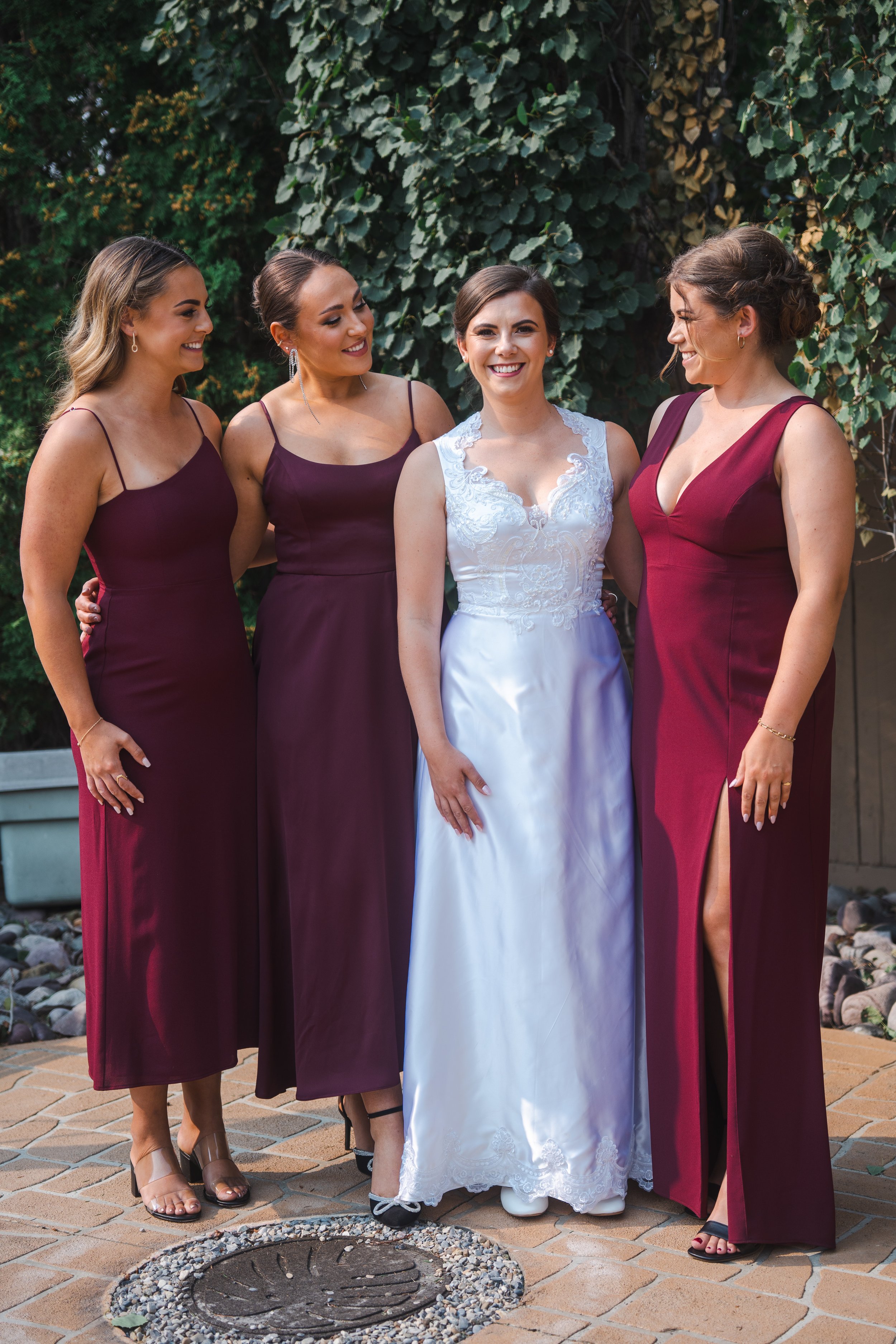 Four women dressed in formal attire standing outdoors on a brick patio, with three wearing matching maroon dresses and one in a white wedding dress, smiling and chatting with each other.