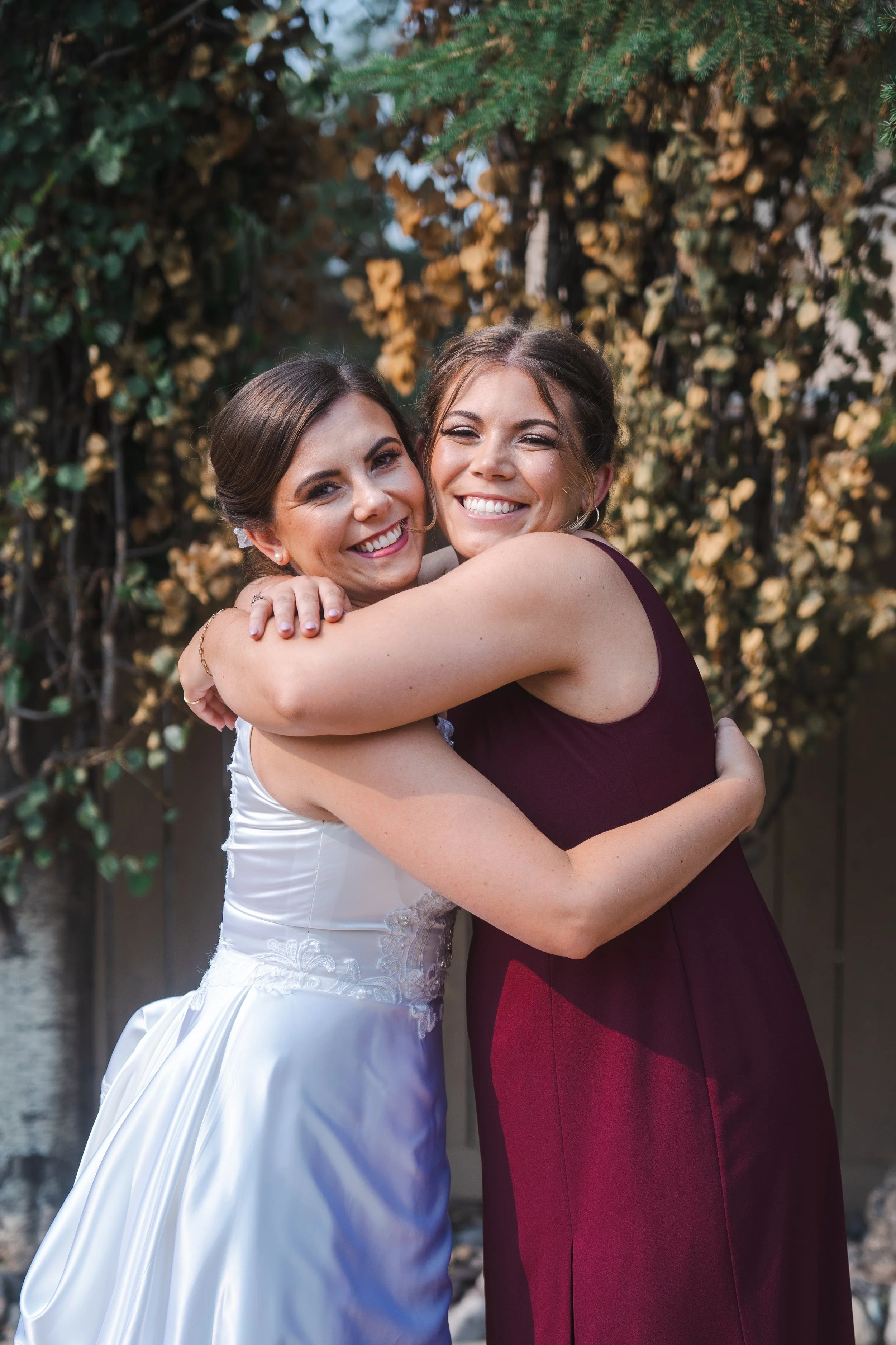 Two women are hugging and smiling happily, one in a white dress and the other in a burgundy dress, standing outdoors with a green leafy background.