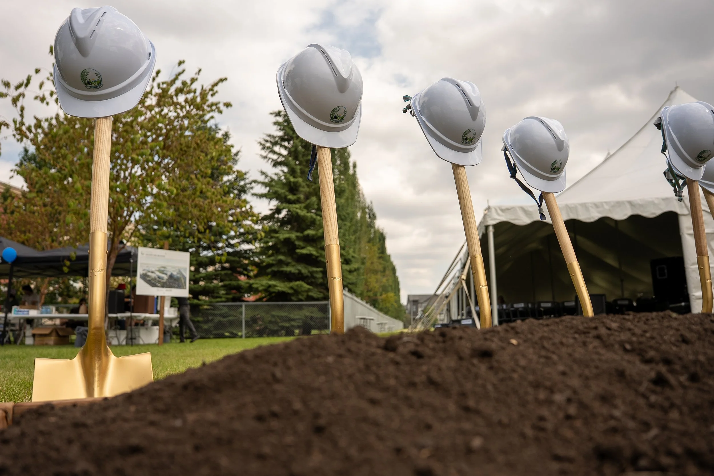 Several white construction helmets with a logo on each, mounted on wooden sticks, are lined up in a row outdoors at a construction site on a cloudy day.