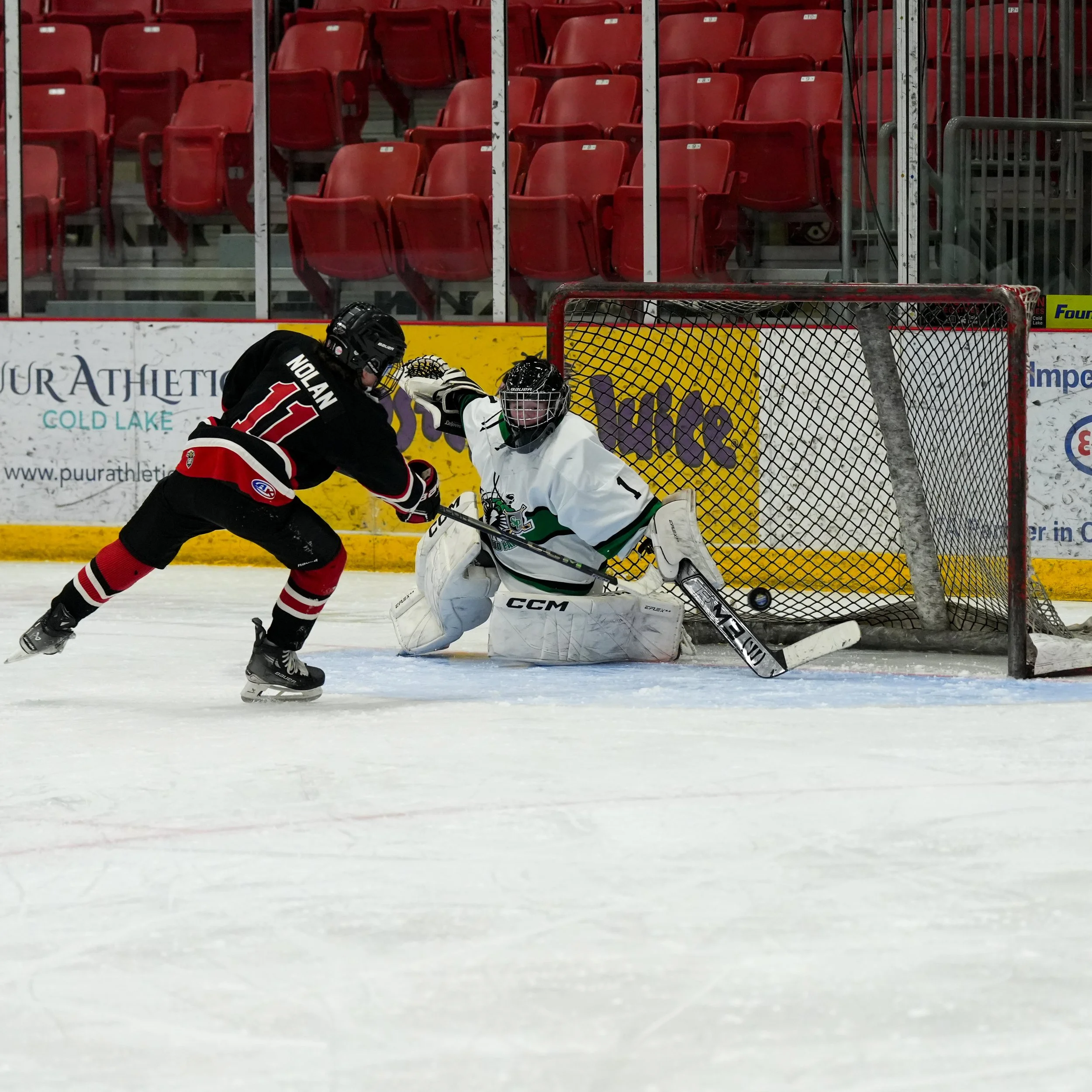 Ice hockey game with players in black and white jerseys near the goal, with rows of red empty seats in the background.