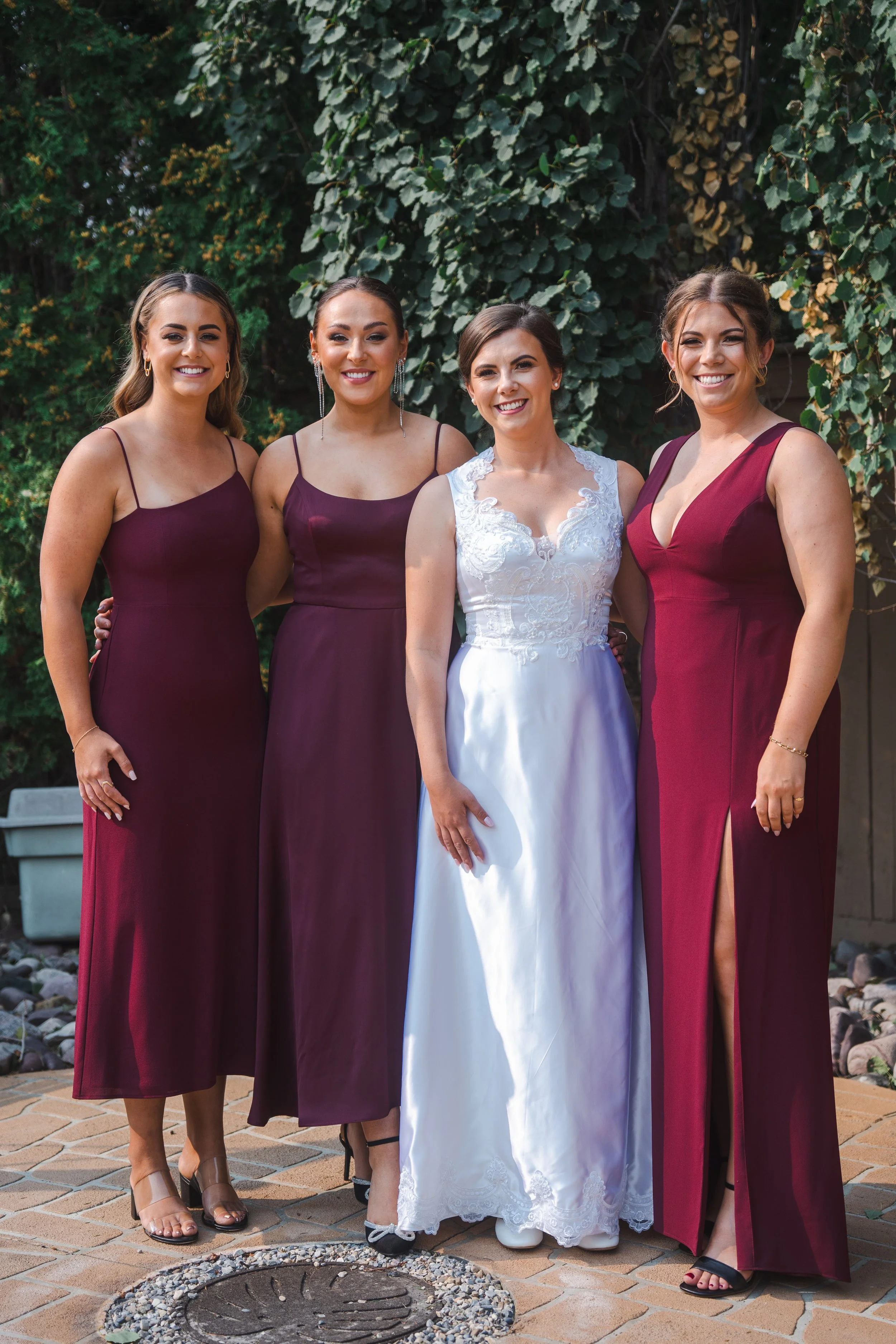Four women standing outdoors in front of green leafy background, dressed in formal attire, smiling, one wearing a white dress, the others wearing burgundy dresses.
