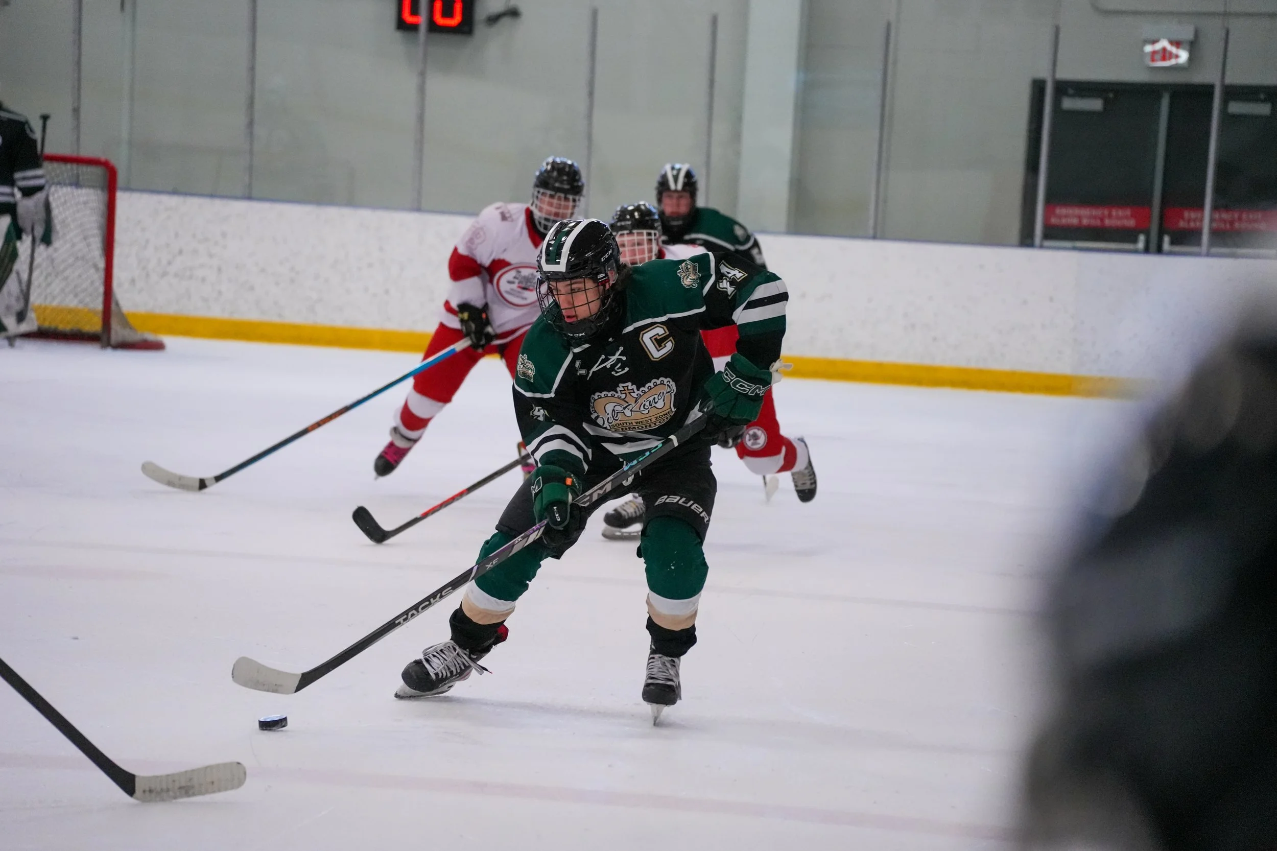 Ice hockey players in a game, with one player in a green jersey with a captain's 'C' and teammates in white and red jerseys, skating on the ice with a puck.