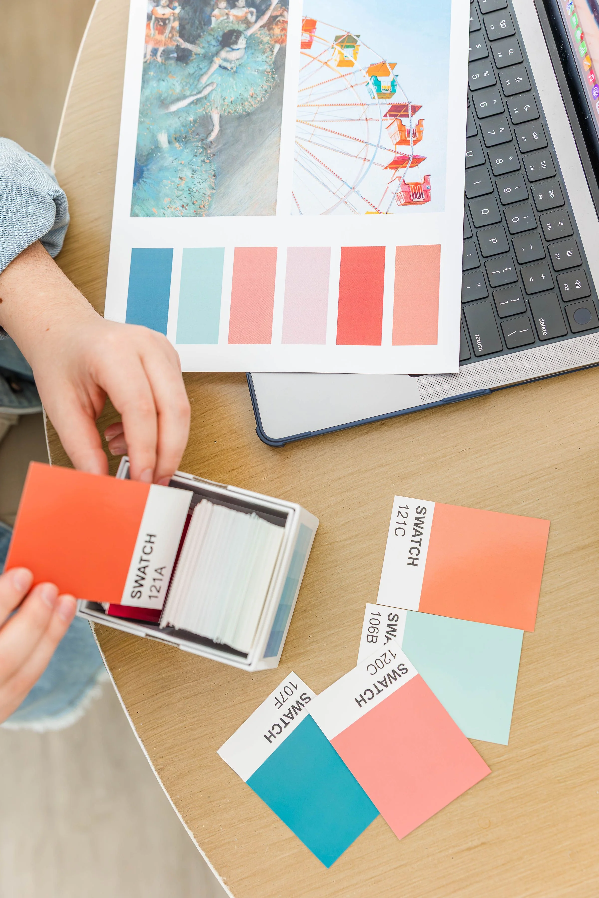 Color swatch samples laid out on a sheet with images of a Ferris wheel and a person swimming in a pool, on a desk next to a laptop with a black keyboard.