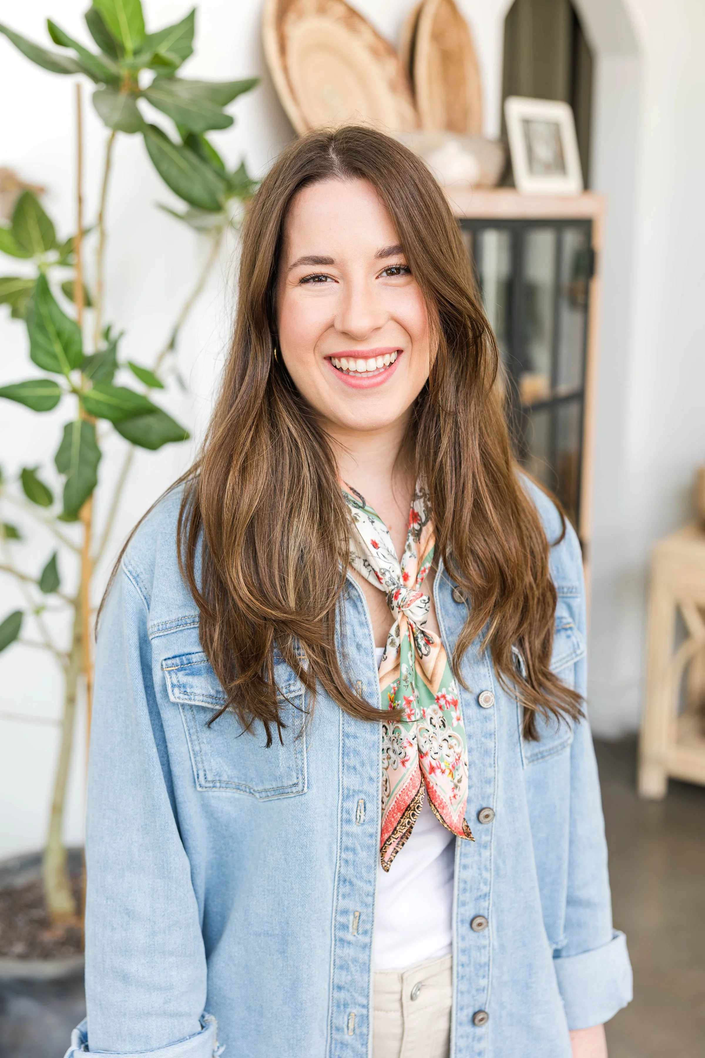 A young woman with long brown hair and a bright smile, wearing a light denim jacket and a colorful scarf, standing indoors near a large plant and a display cabinet with decor items.