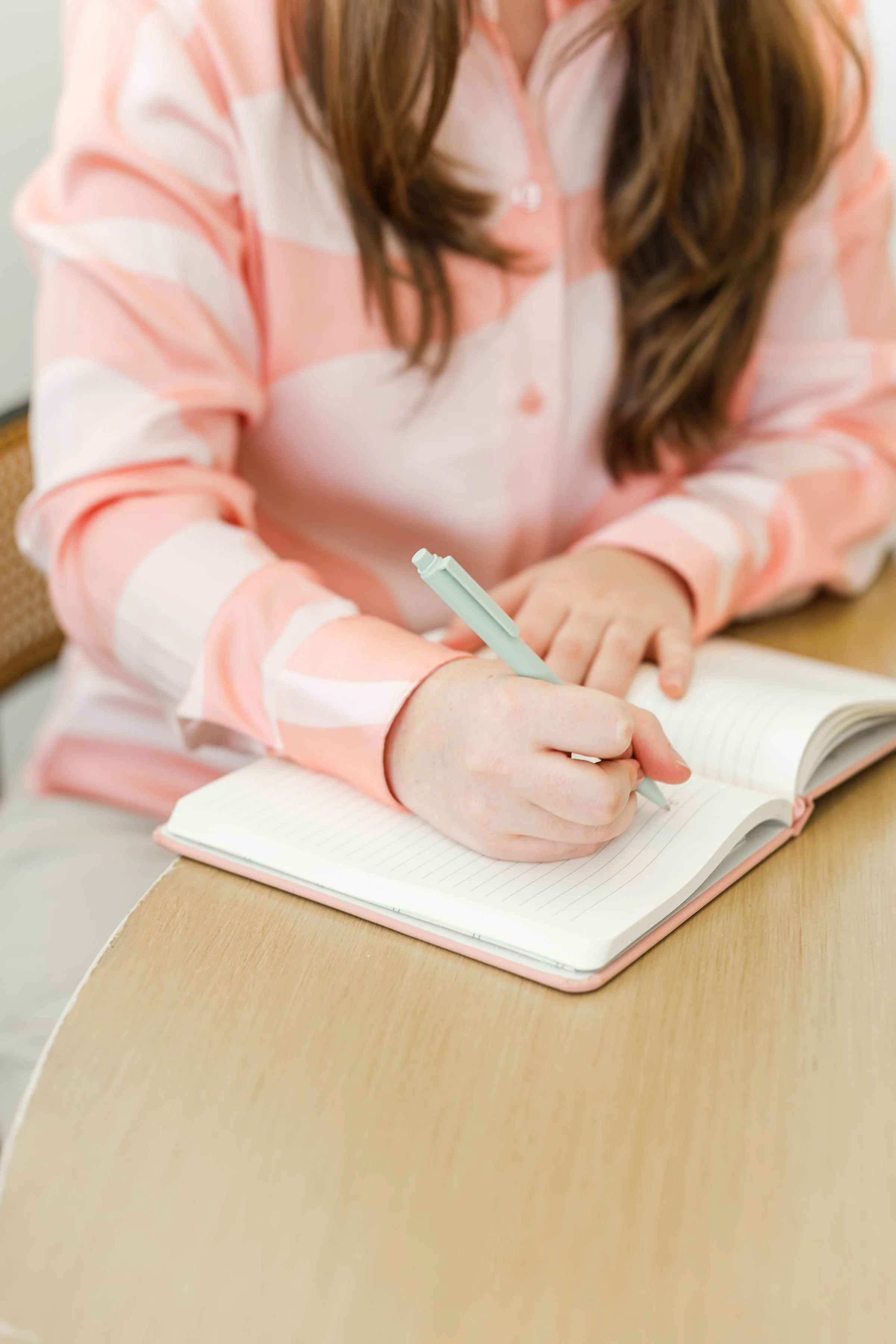 A woman with brown hair, wearing a pink and white striped shirt, writing in a lined pink notebook with a light blue pen while sitting at a wooden table.