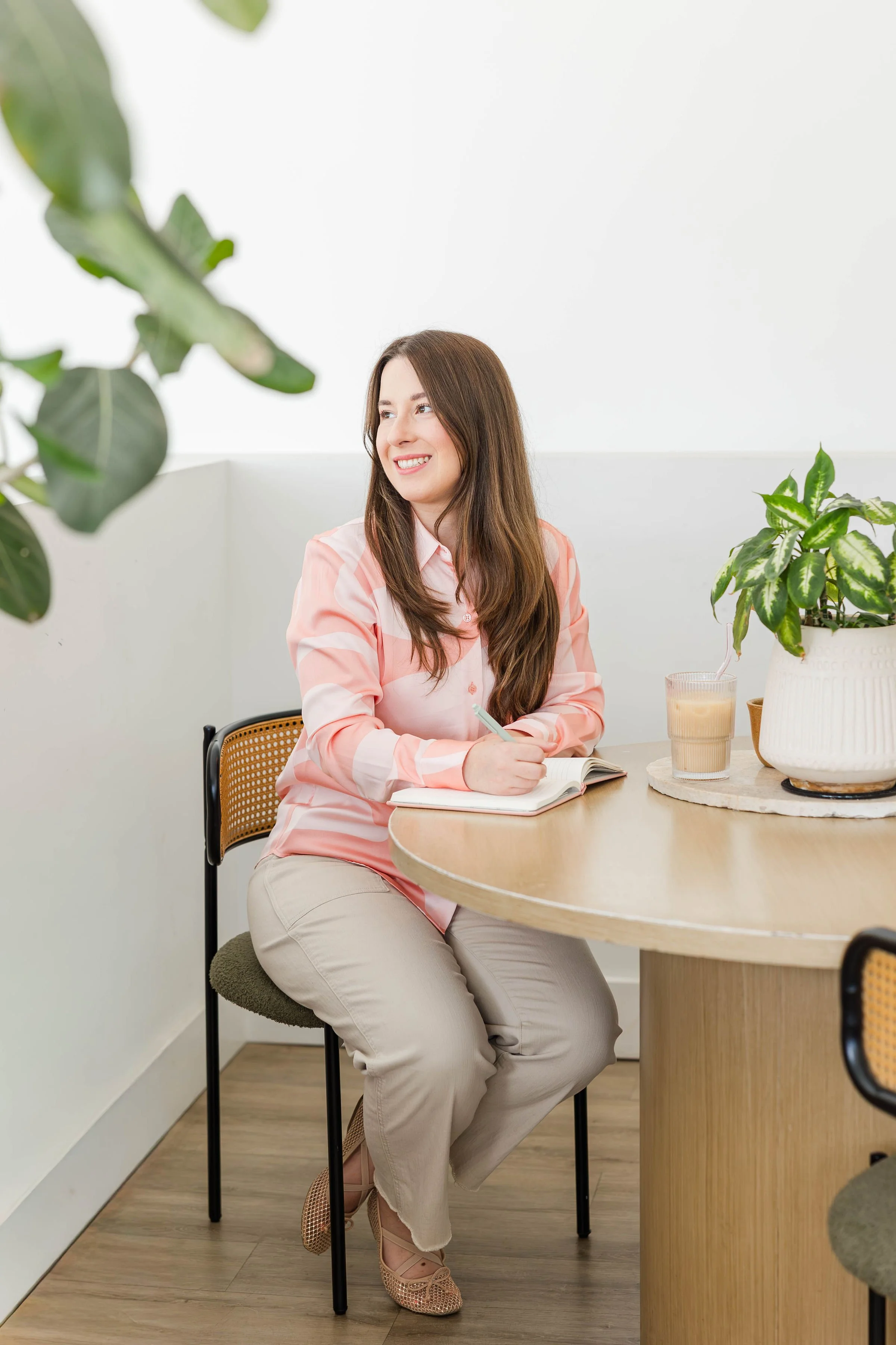 A woman with long brown hair, wearing a pink and white striped shirt and beige pants, sitting at a round wooden table with a notebook and a pen, smiling and looking to her left. There are green plants nearby, one on the table in a white pot and the other partially visible in the foreground.