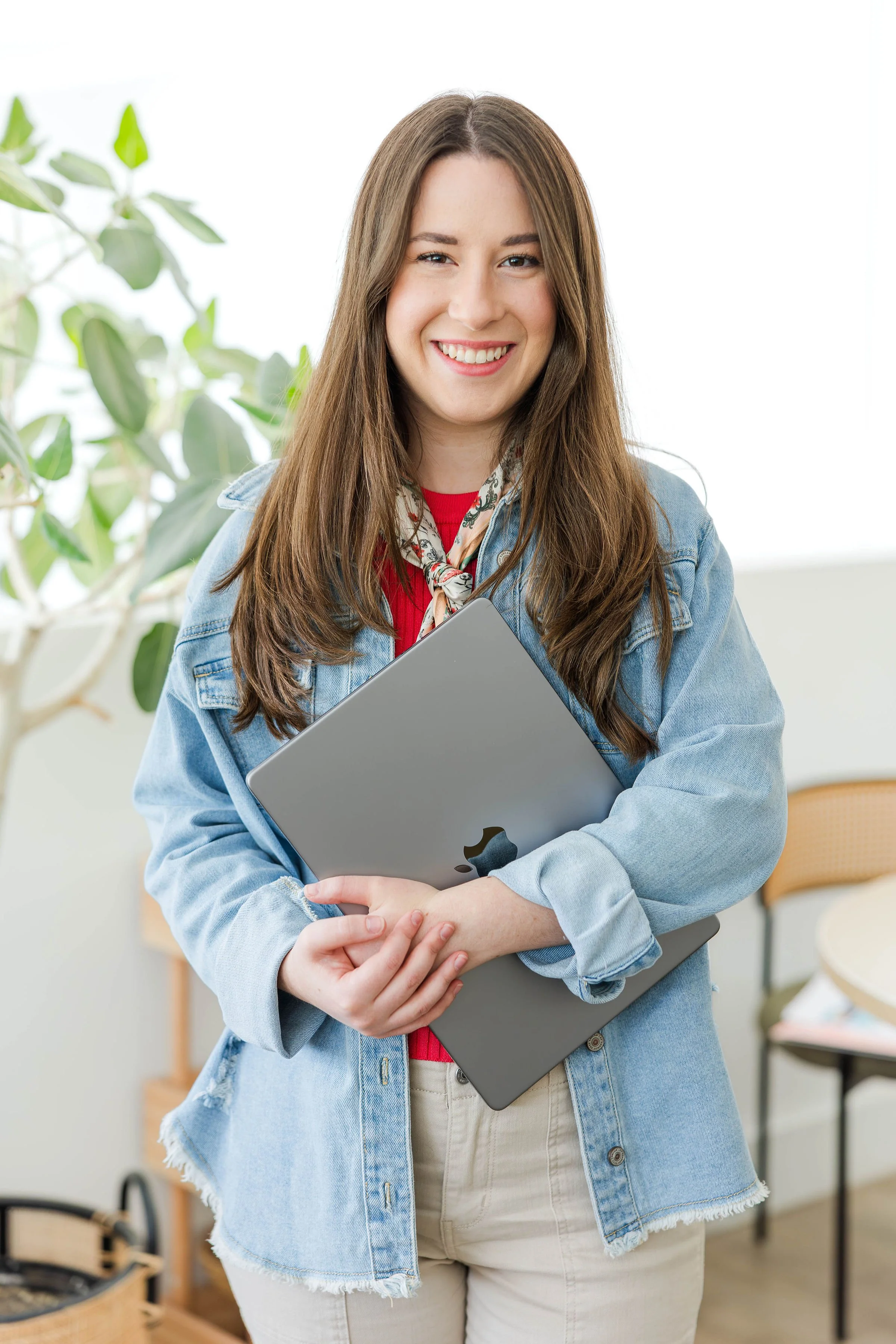 A young woman with long brown hair wearing a denim jacket and scarf, holding a silver laptop, smiling in a bright indoor space with green plants and chairs.