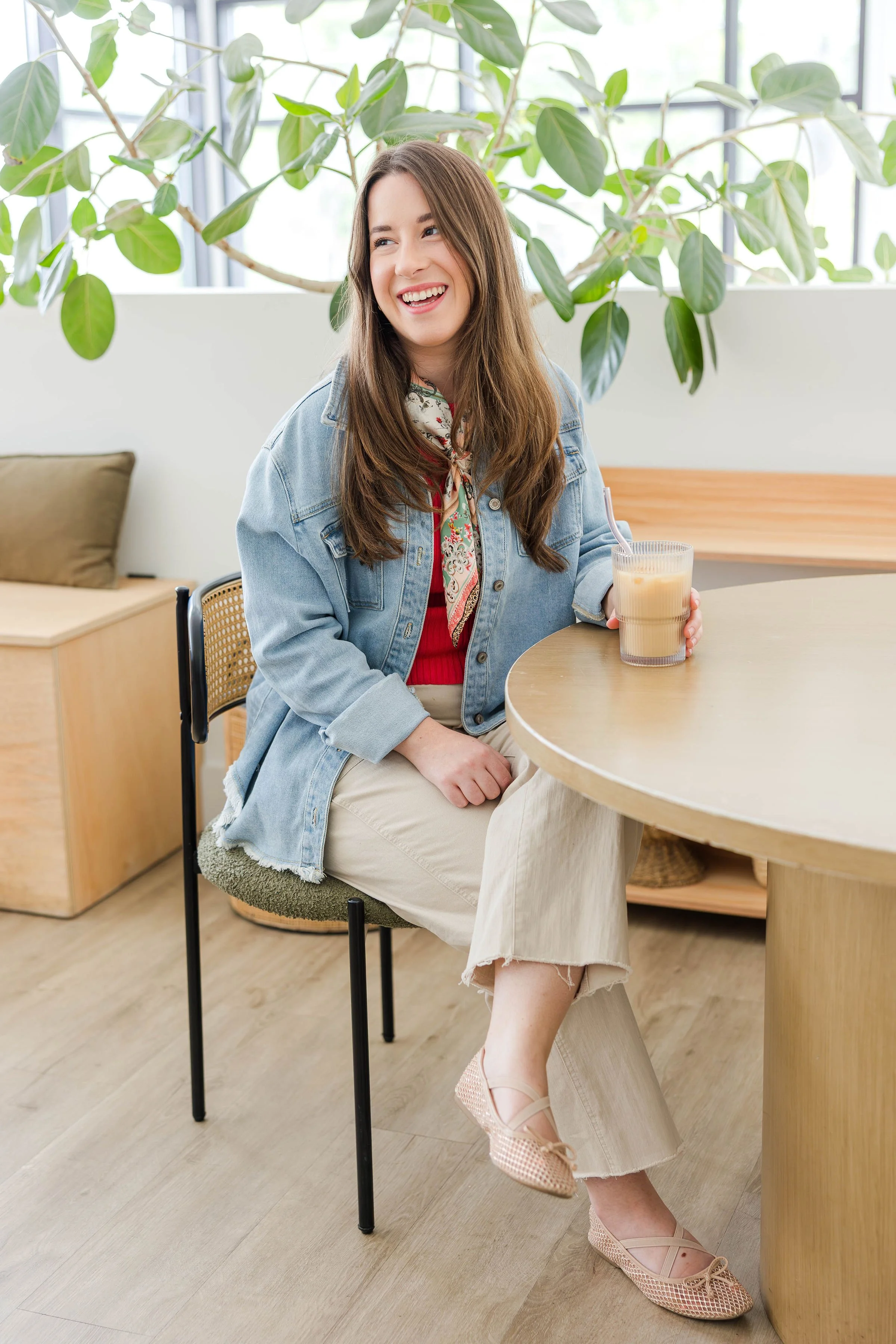 A young woman with long brown hair, wearing a light denim jacket, red top, beige pants, and flats, smiling and sitting at a round wooden table with a glass of iced coffee. Behind her, there are large green plants and a window with natural light.
