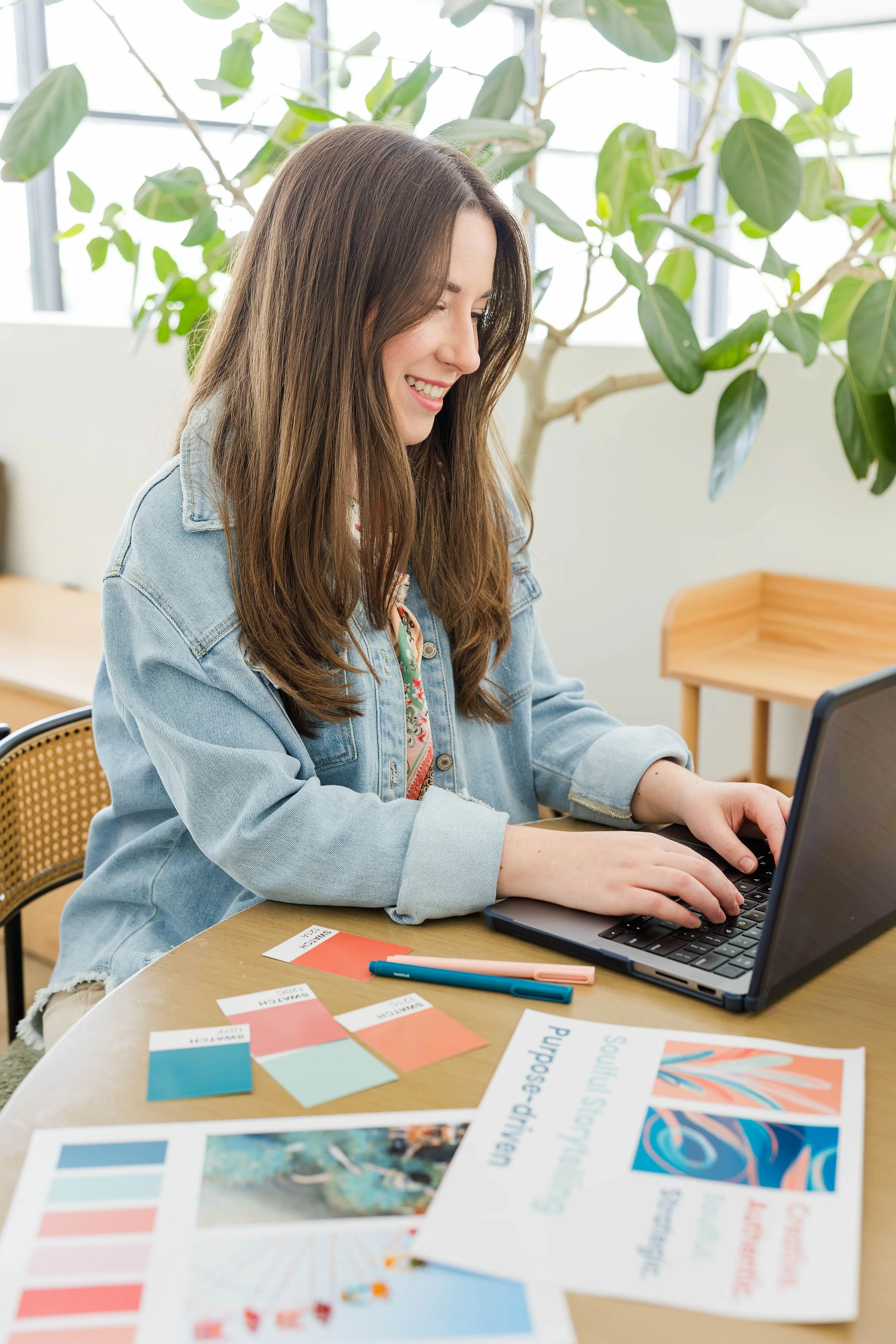 A young woman with long brown hair and a denim jacket smiling while working on a laptop at a desk in a bright room with large green plants in the background.