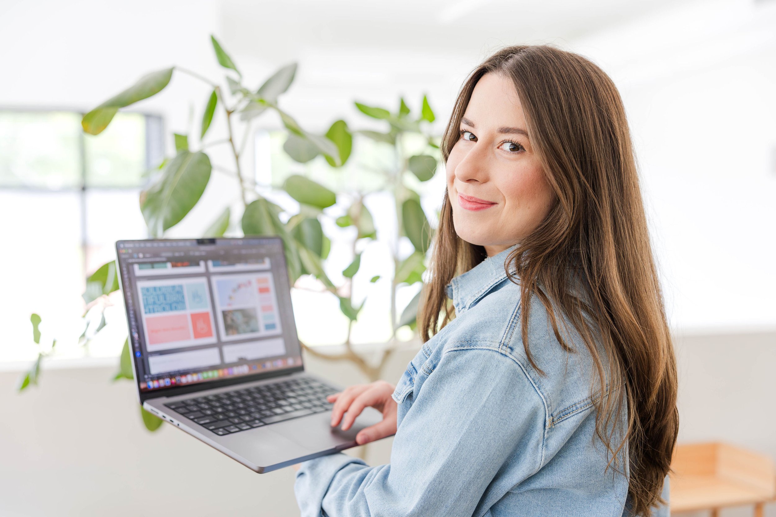 A young woman with long brown hair holding a laptop working on graphic designs in a bright room with a plant in the background, smiling at the camera.