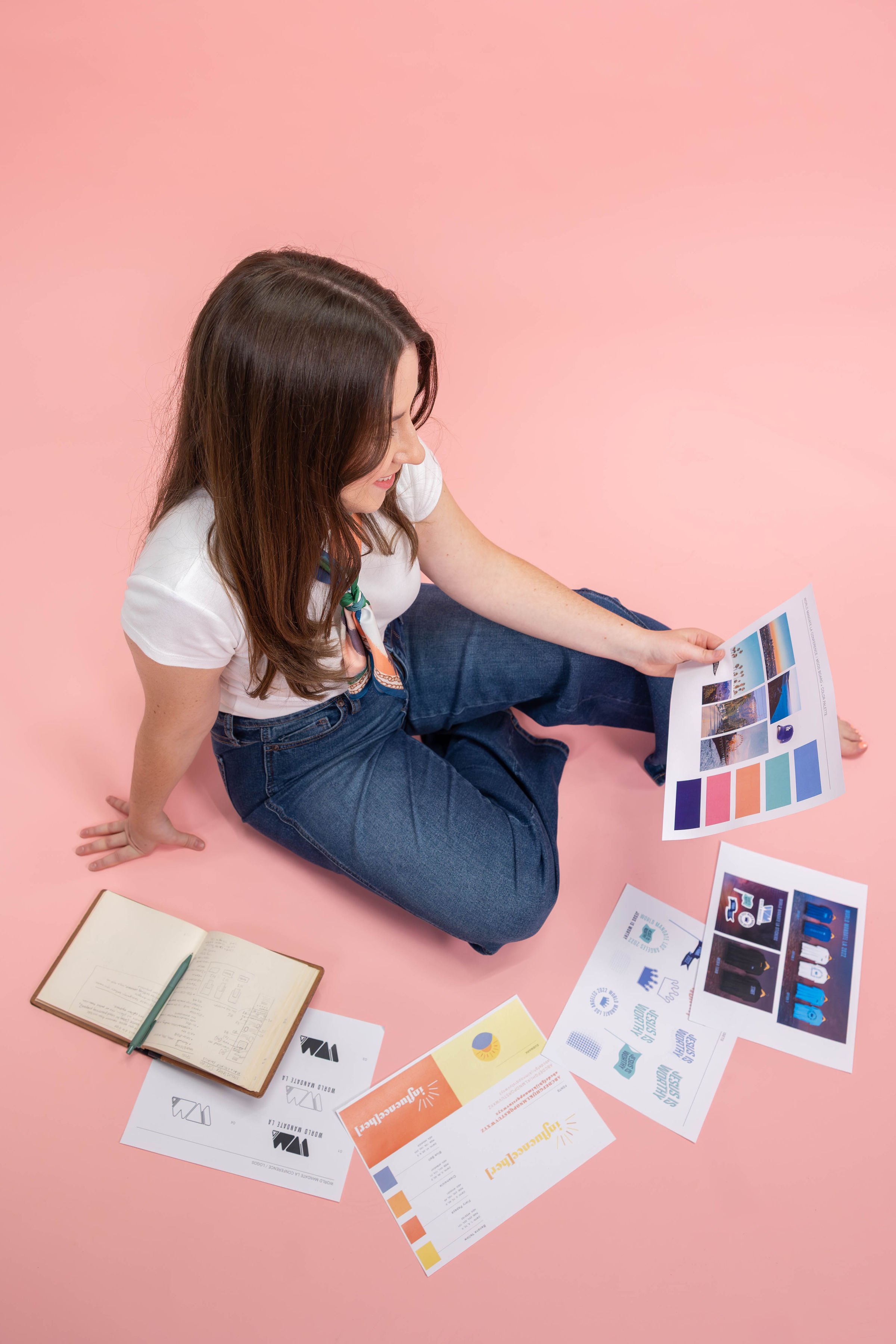 A woman sitting on a pink floor, looking at printed flyers and brochures related to branding and marketing. An open notebook with handwritten notes is nearby.