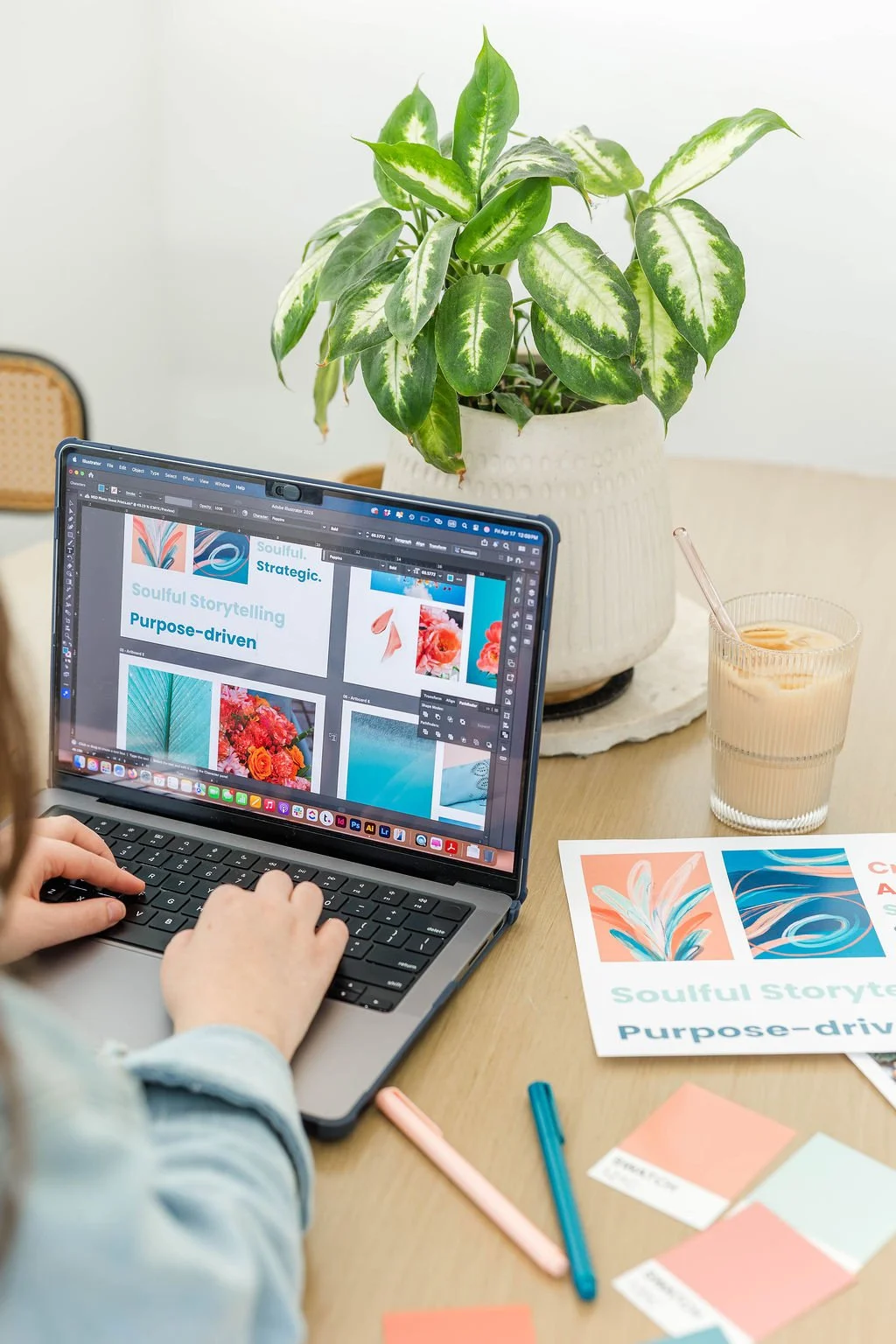 A person working on a laptop with design project images on the screen. There is a potted green plant, a glass of iced coffee, and printed color swatches on the wooden desk.