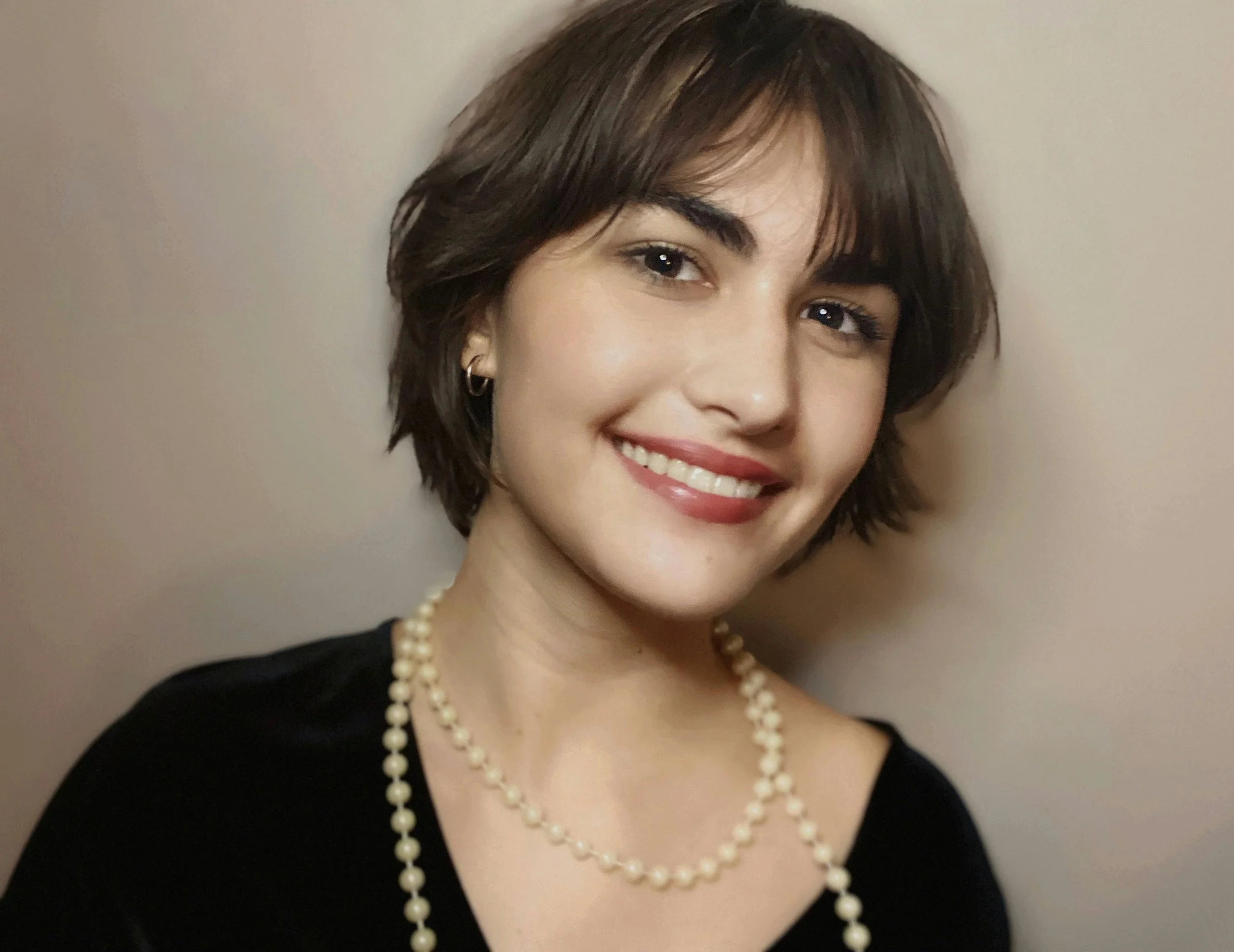 A young woman with short brown hair, wearing a black top, pearl necklace, and earrings, smiling and looking at the camera against a plain background.