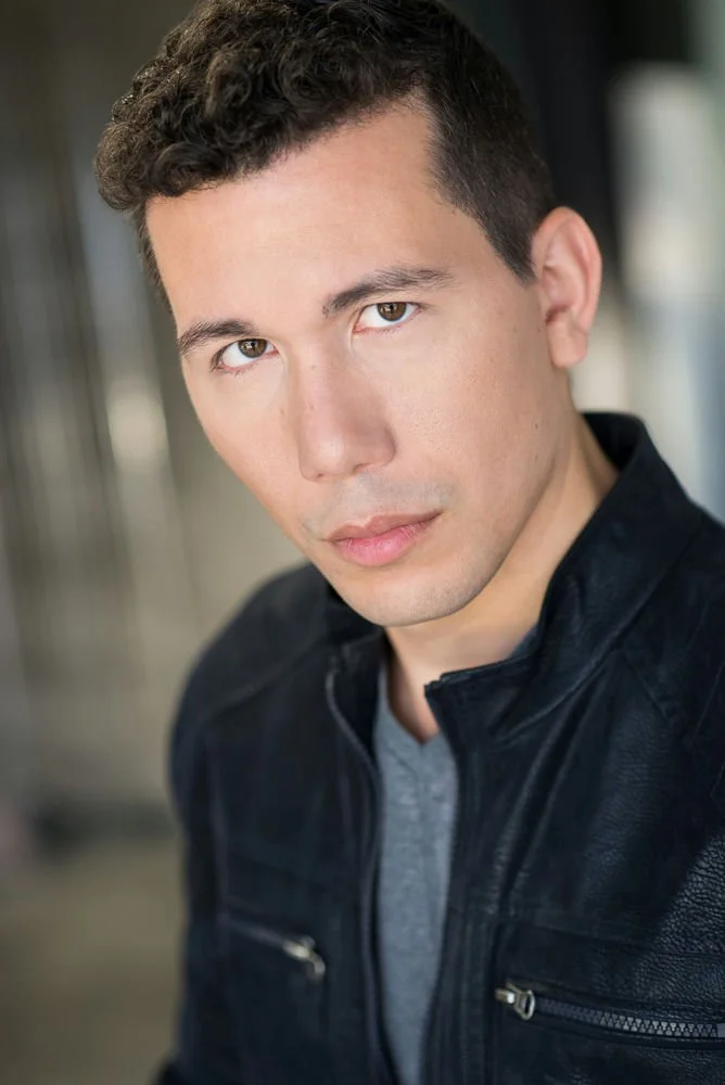 A young woman with curly reddish-brown hair and brown eyes, wearing a black lace top and layered gold necklaces, smiling at the camera.