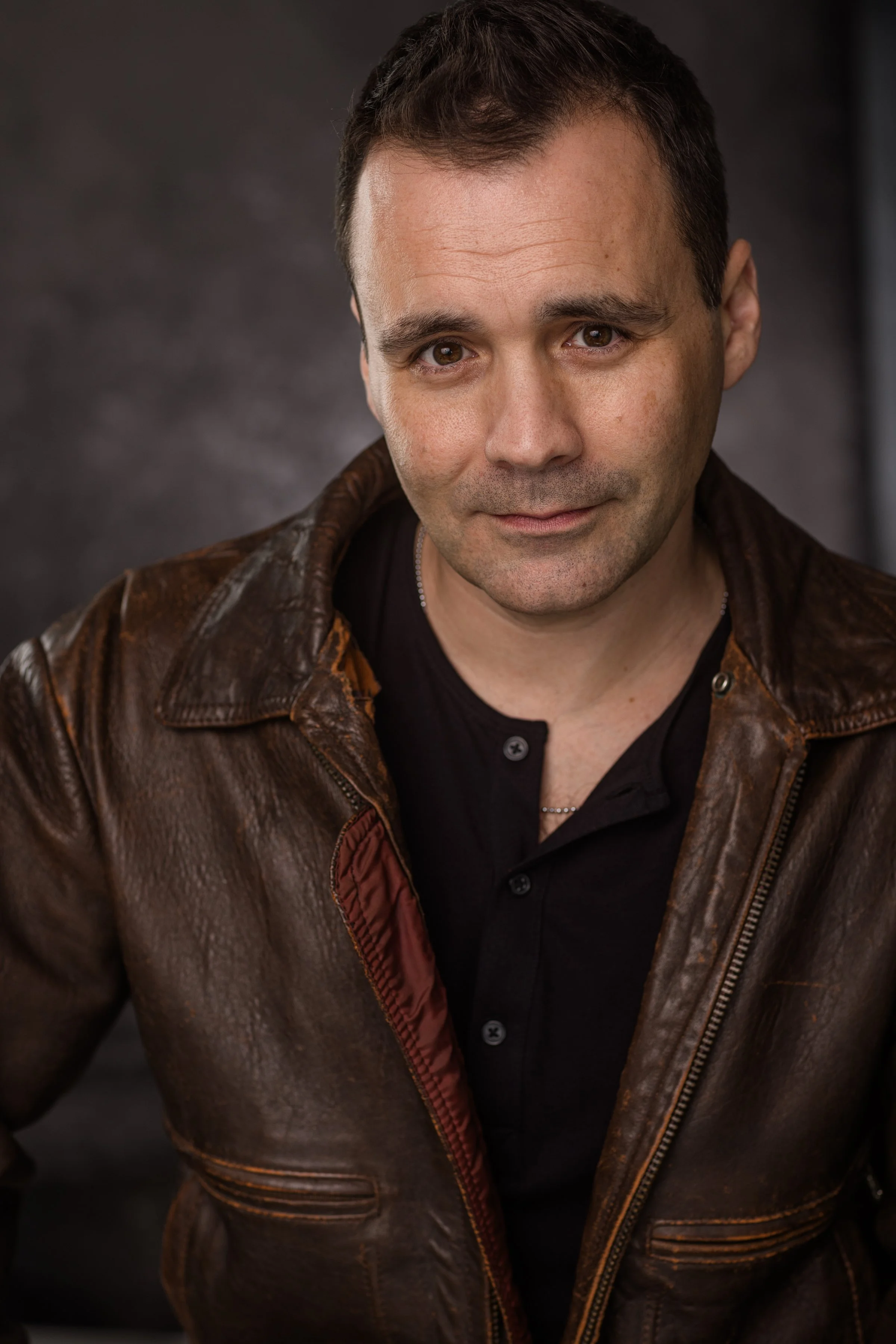 Close-up portrait of a woman with dark brown, textured dreadlocks, light makeup, and wearing a dark green top, against a neutral background.