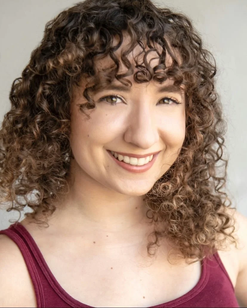 A smiling woman with dark curly hair, wearing hoop earrings and a cream-colored sweater, against a dark background.