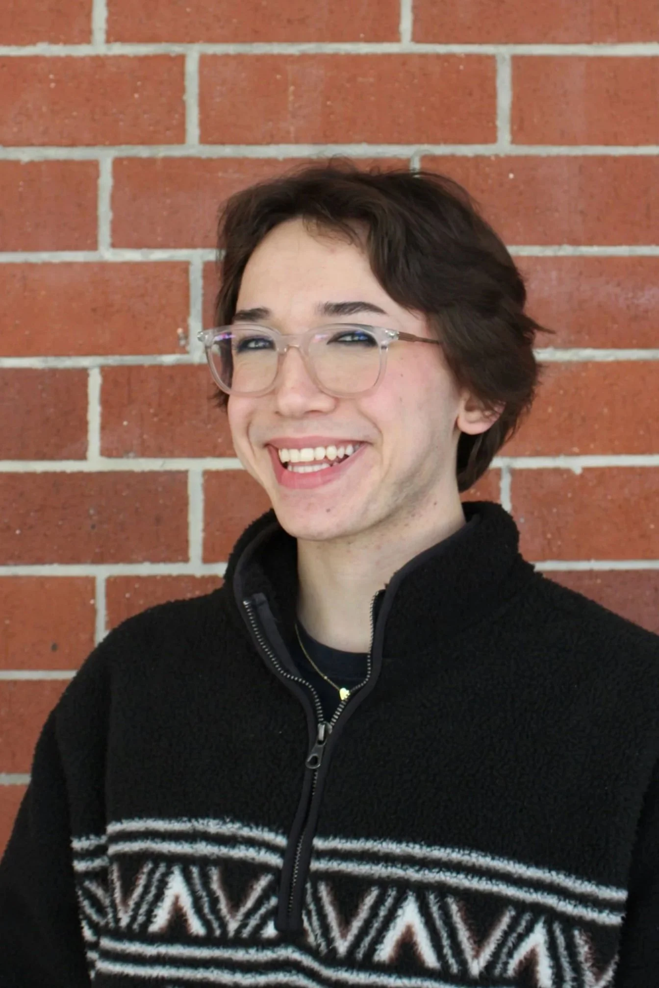 Young woman with curly hair standing in front of a brick wall, wearing a black shirt, looking to the side, adjusting her hair.