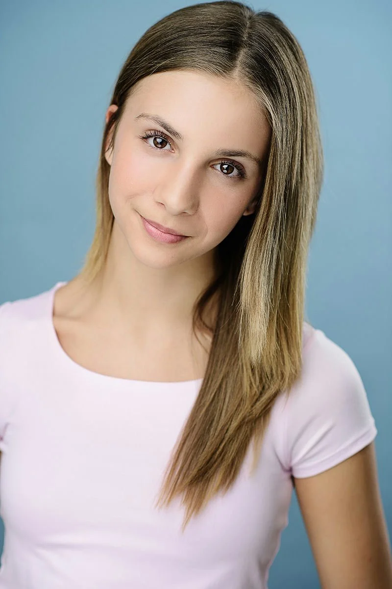Young man with curly dark hair, smiling, wearing a brown jacket and white t-shirt, standing outdoors in front of green leafy bushes.