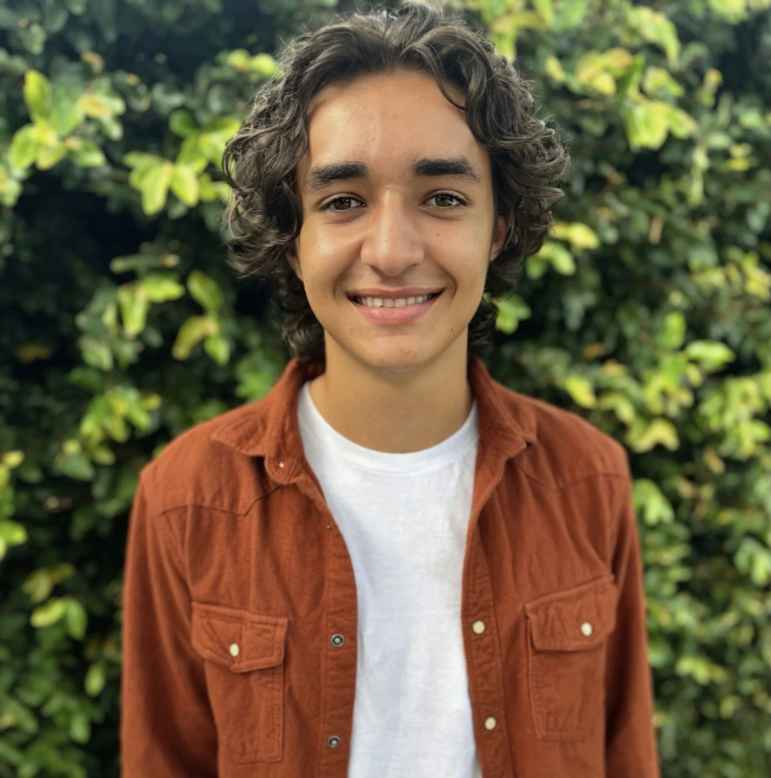 Young man with curly dark hair, smiling, wearing a brown jacket and white t-shirt, standing outdoors in front of green leafy bushes.