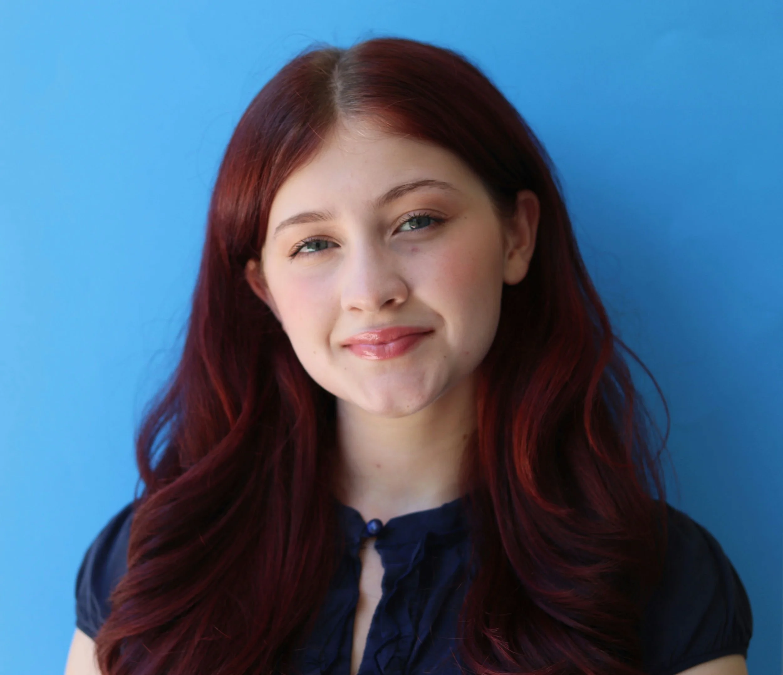 Portrait of a young woman with red wavy hair and blue eyes, wearing a dark blue blouse, standing against a blue background.