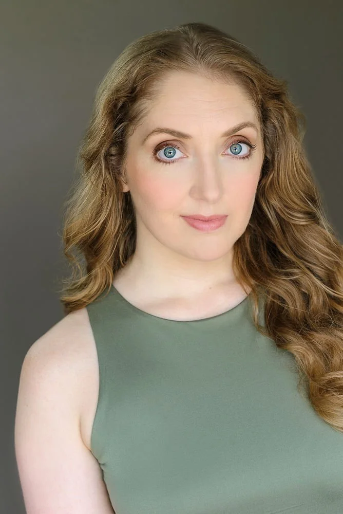 A woman with curly red hair wearing a sleeveless red top, looking at the camera with a slight smile.