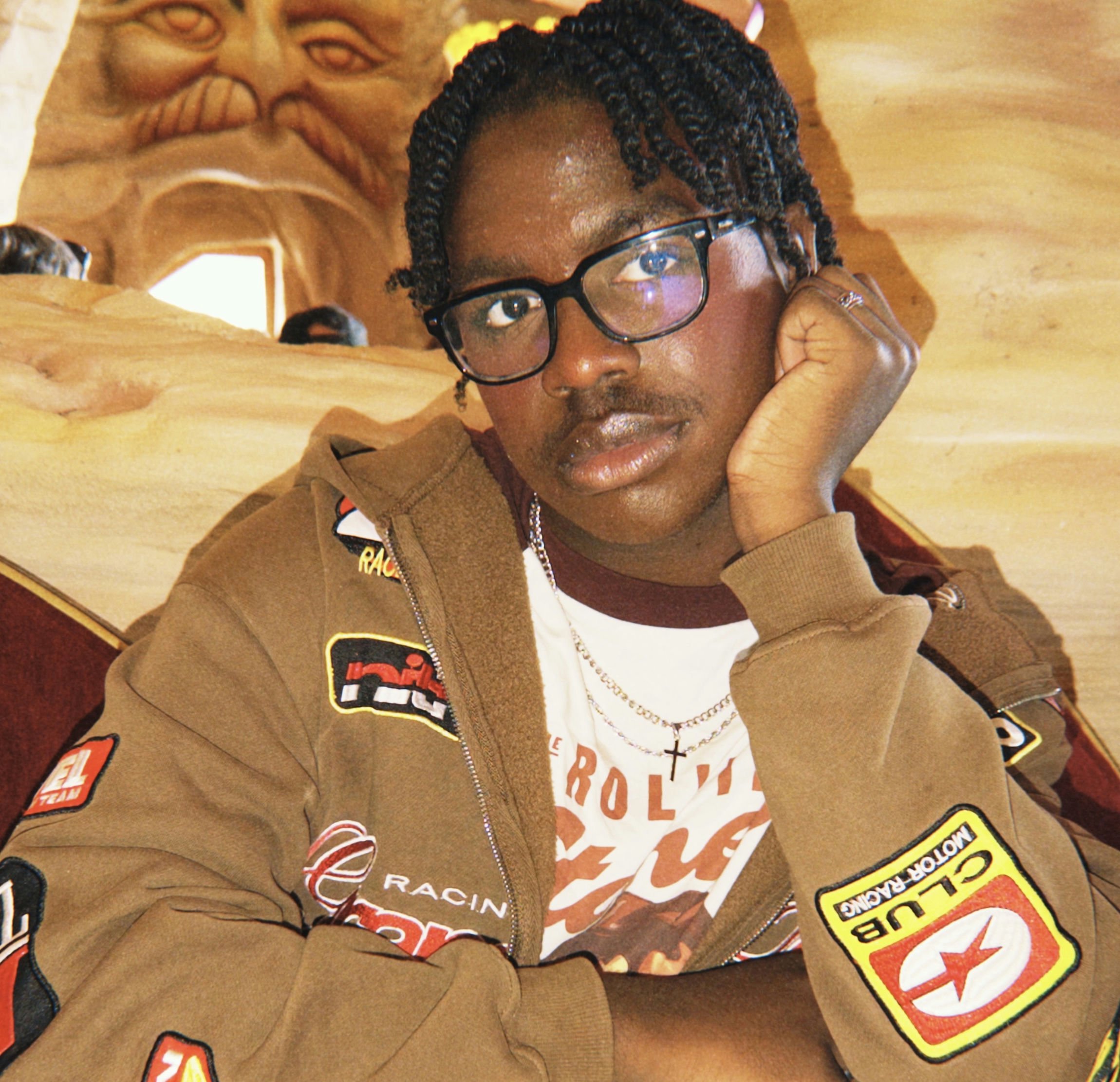 Close-up portrait of a young person with curly dark hair and green eyes, wearing a white collared shirt, standing indoors with blurred windows in the background.