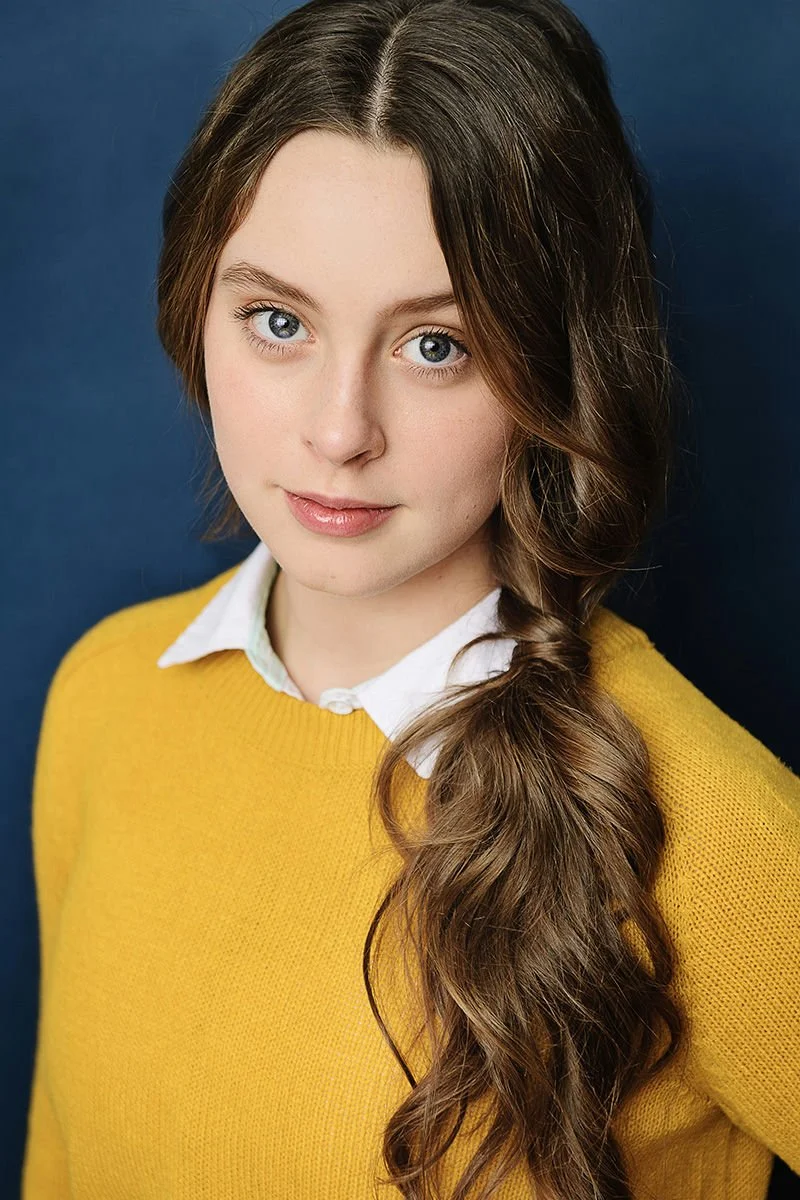A young person with dark hair, neutral expression, wearing a dark zip-up hoodie, looking at the camera against a dark background.