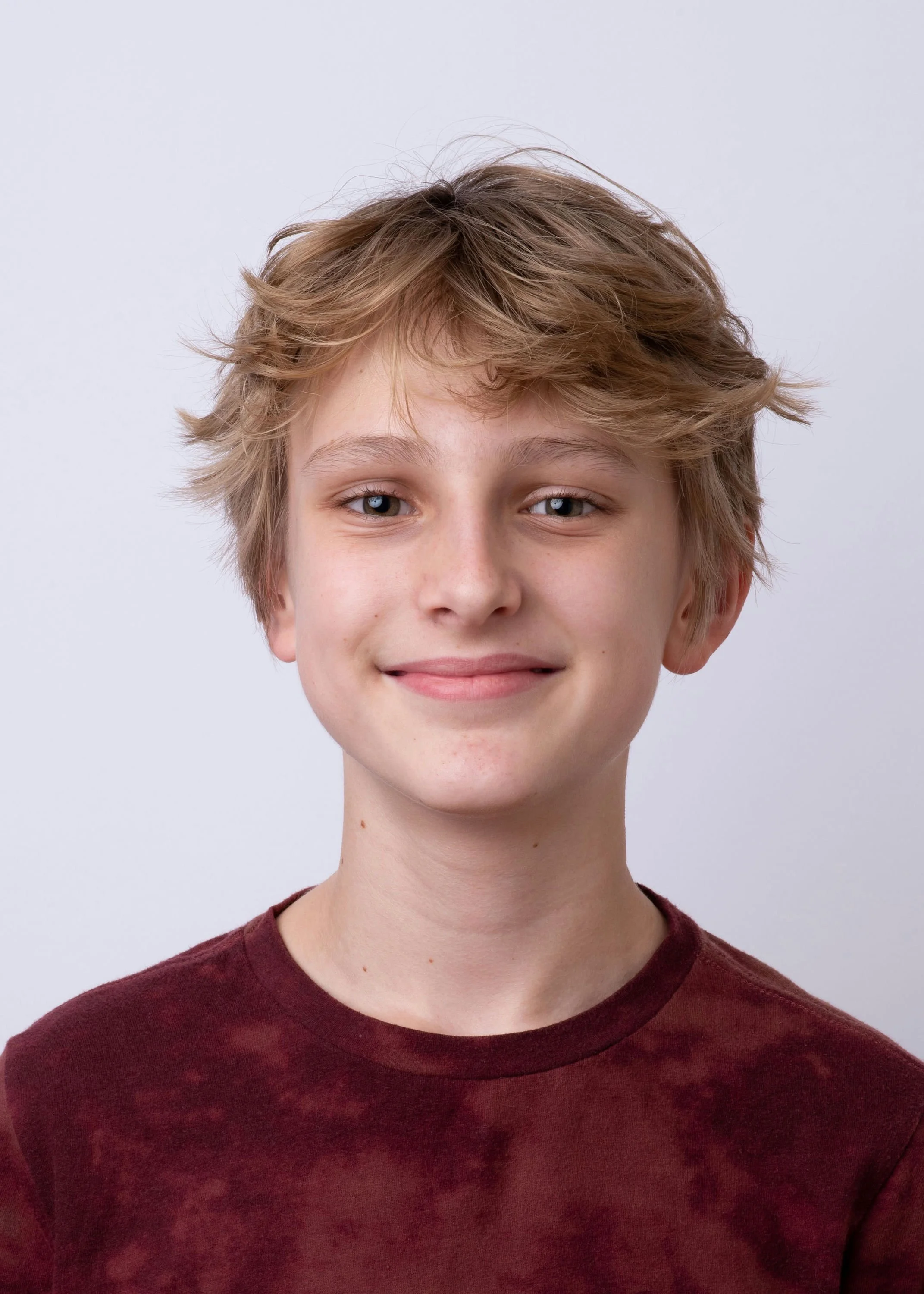 A young boy with tousled blond hair, wearing a maroon shirt, smiling at the camera against a plain light gray background.