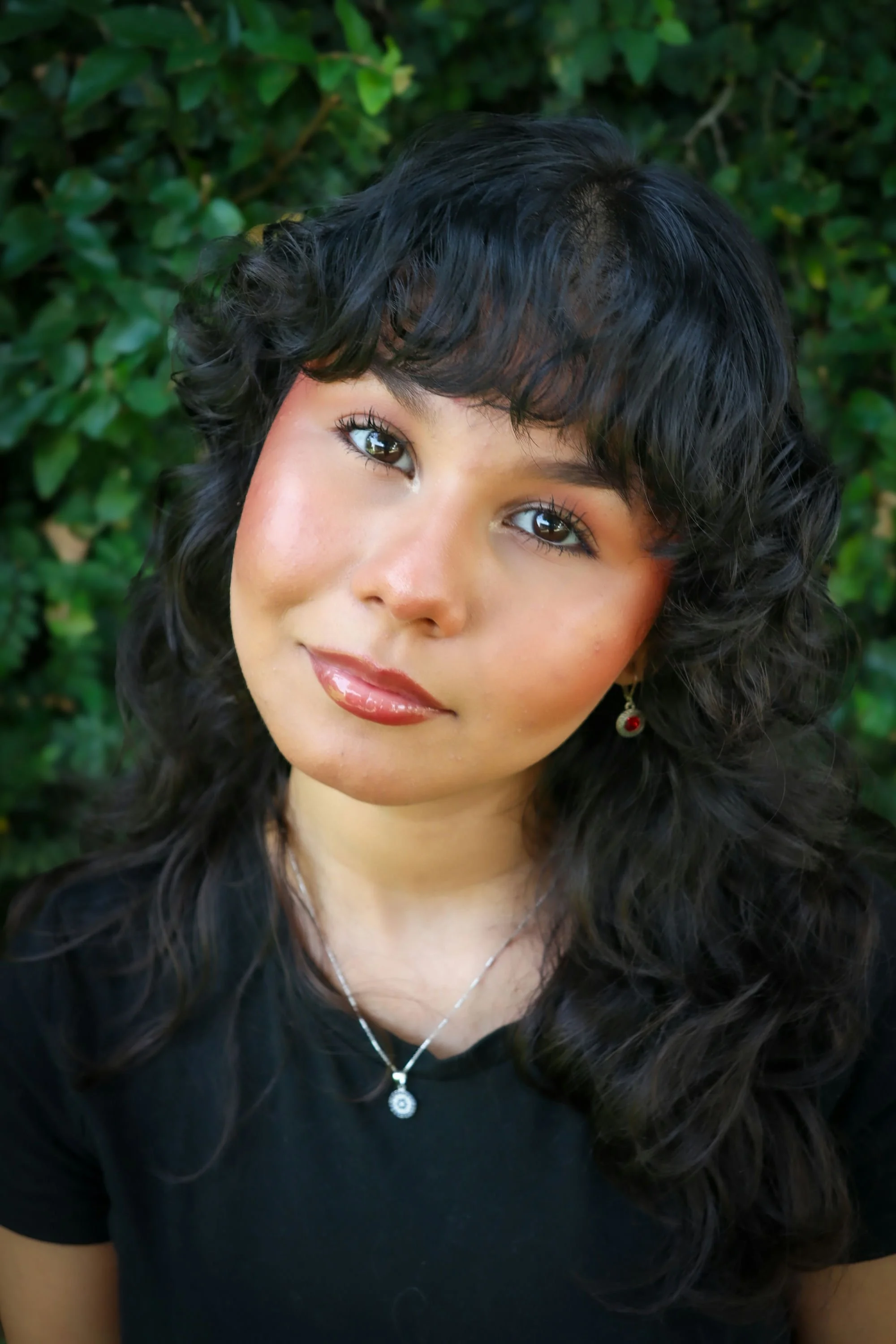Close-up of a young woman with shoulder-length brown hair, wearing a silver studded headband, hoop earrings, and a white t-shirt, smiling slightly against a plain wall background.
