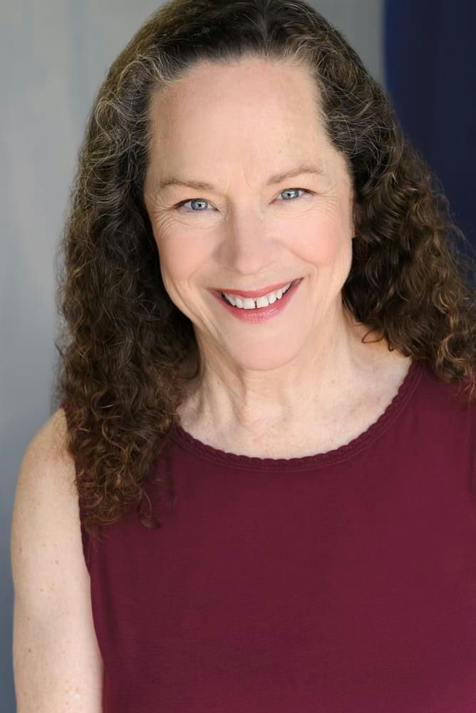 A smiling woman with dark curly hair, wearing hoop earrings and a cream-colored sweater, against a dark background.