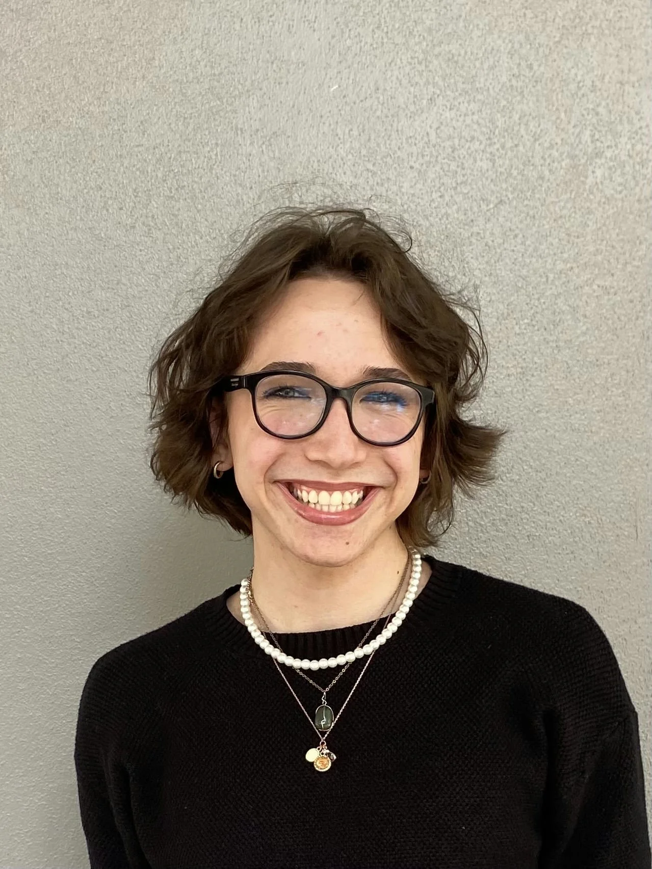 A smiling woman with short, wavy brown hair wearing glasses, a black sweater, a pearl necklace, and layered necklaces, standing against a plain beige wall.