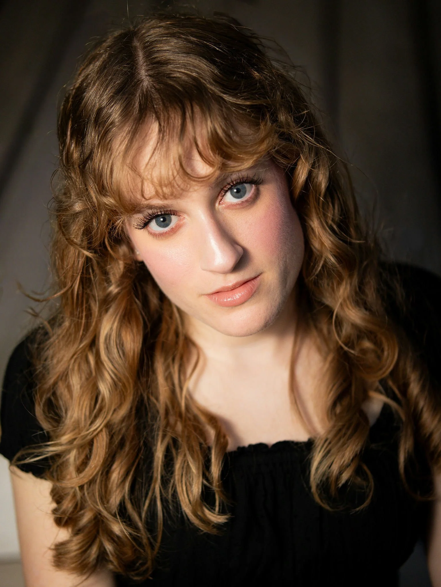 A smiling young person with short dark hair and freckles, wearing a light gray button-up shirt over a white t-shirt, posing against a black background.