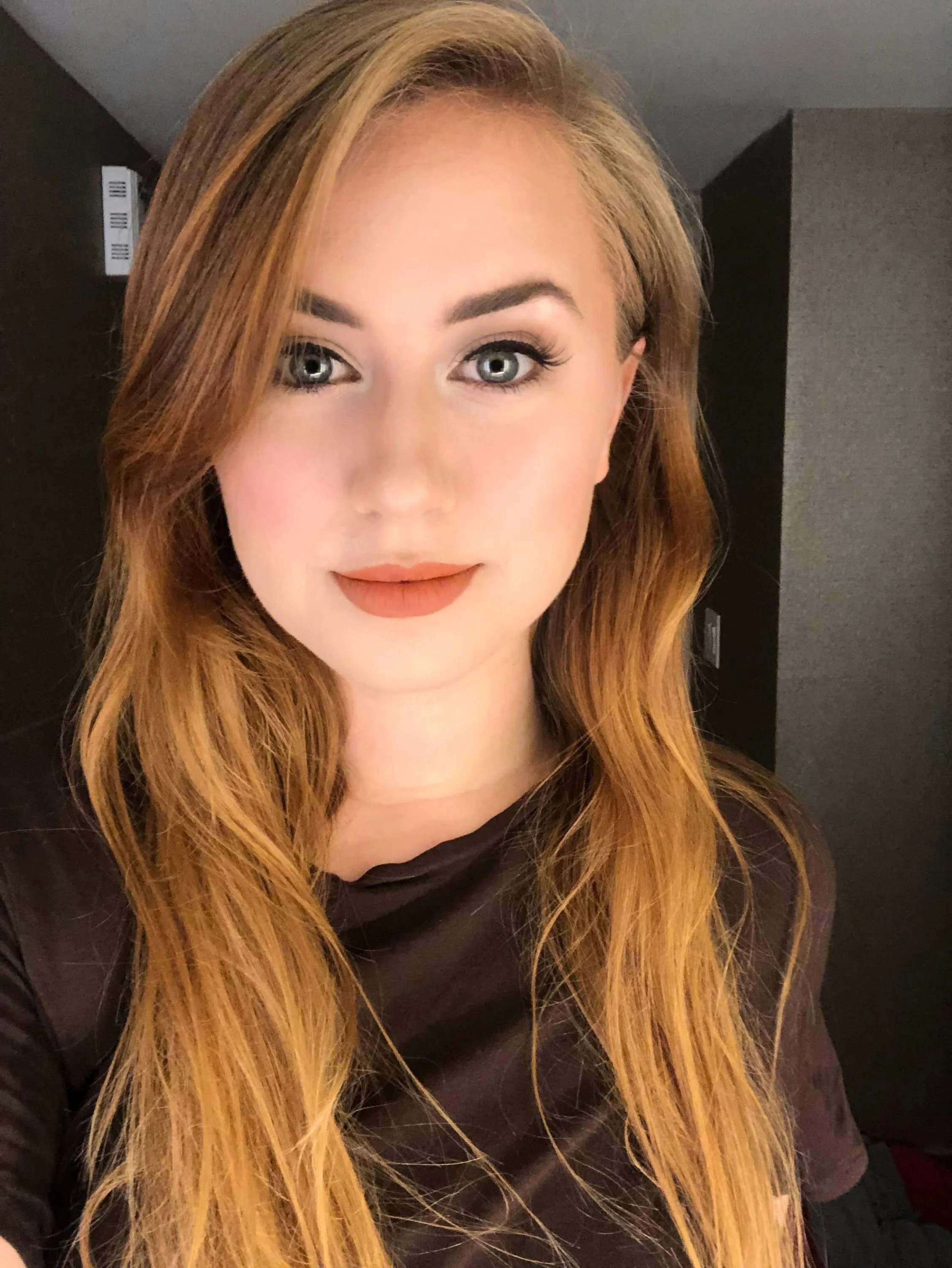 A young woman with curly reddish-brown hair and brown eyes, wearing a black lace top and layered gold necklaces, smiling at the camera.