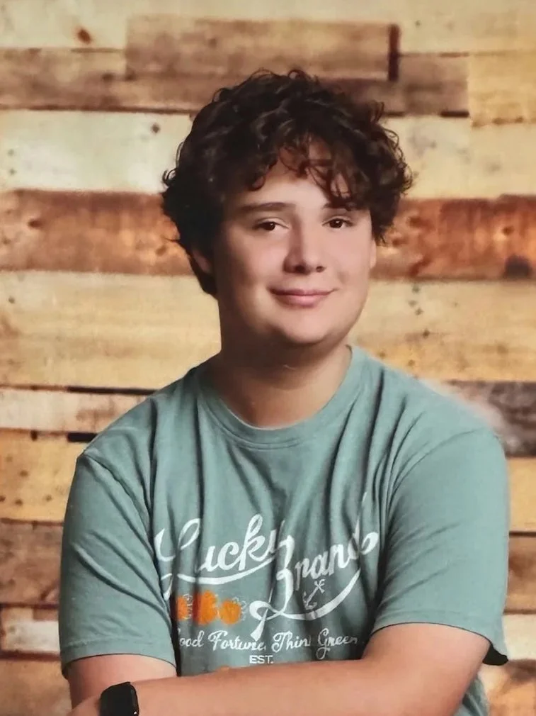 Close-up portrait of a young man with dark wavy hair, light skin, and wearing a gray collared shirt, looking directly at the camera against a plain white background.