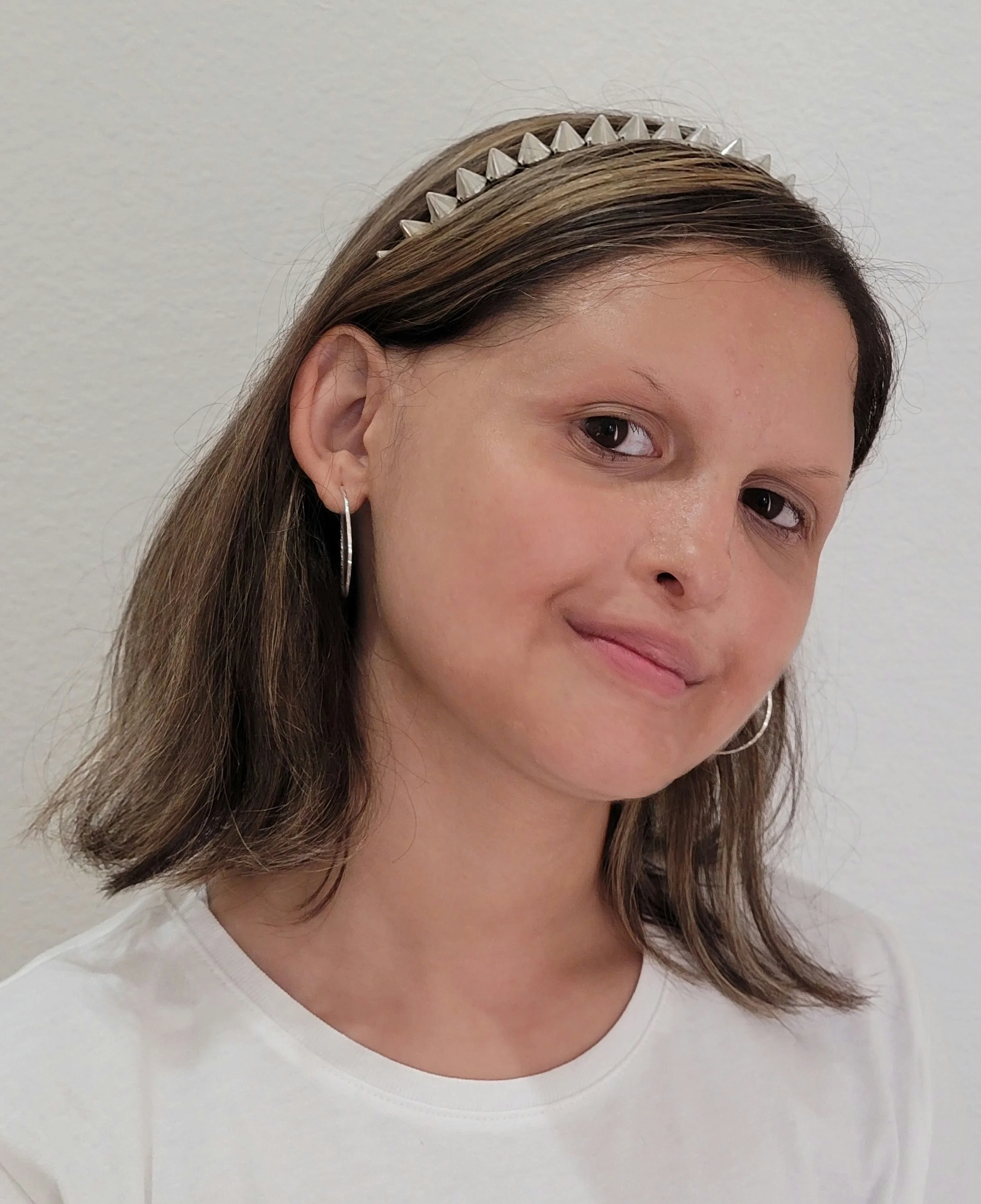 Close-up of a young woman with shoulder-length brown hair, wearing a silver studded headband, hoop earrings, and a white t-shirt, smiling slightly against a plain wall background.