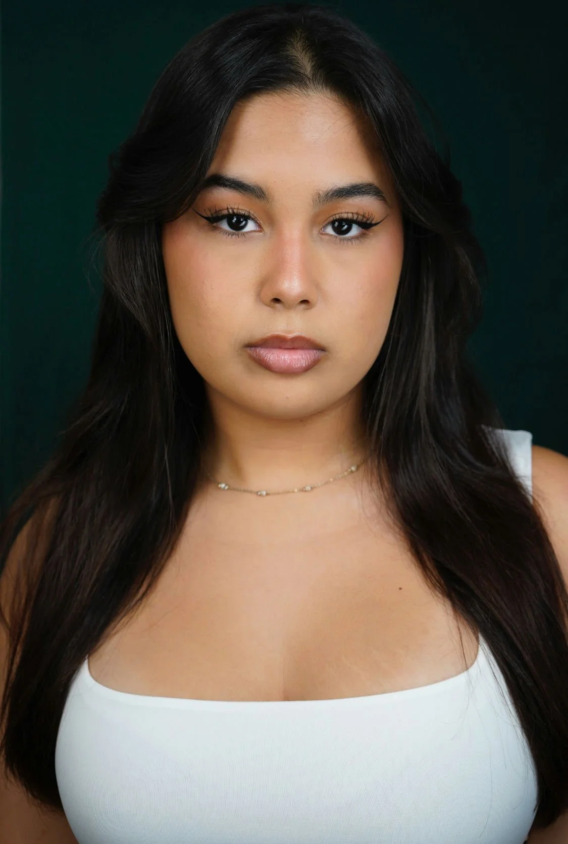 Close-up portrait of a woman with dark brown, textured dreadlocks, light makeup, and wearing a dark green top, against a neutral background.