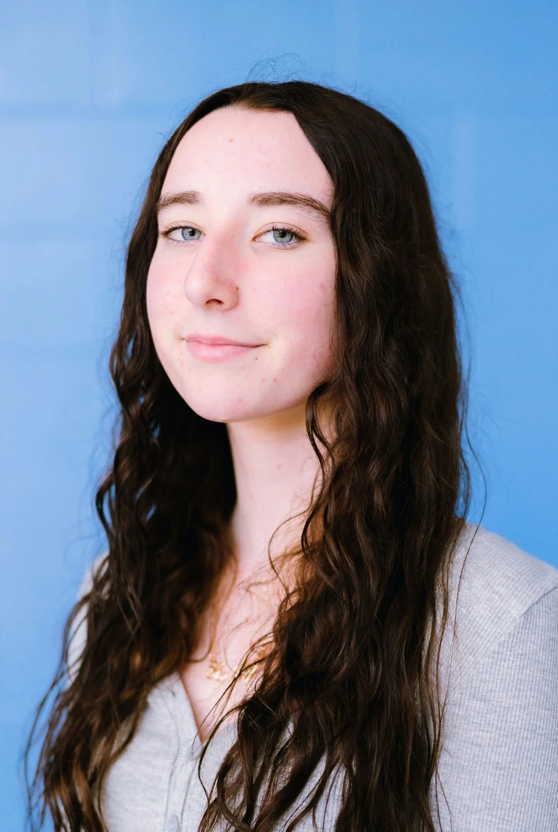 Portrait of a young woman with long, wavy brown hair, blue eyes, fair skin, and a slight smile, wearing a light gray top, against a blue background.