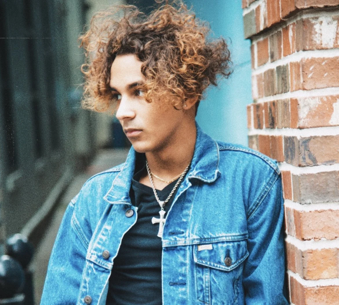 Portrait of a person with long dreadlocks, wearing a dark blue shirt, against a dark background.