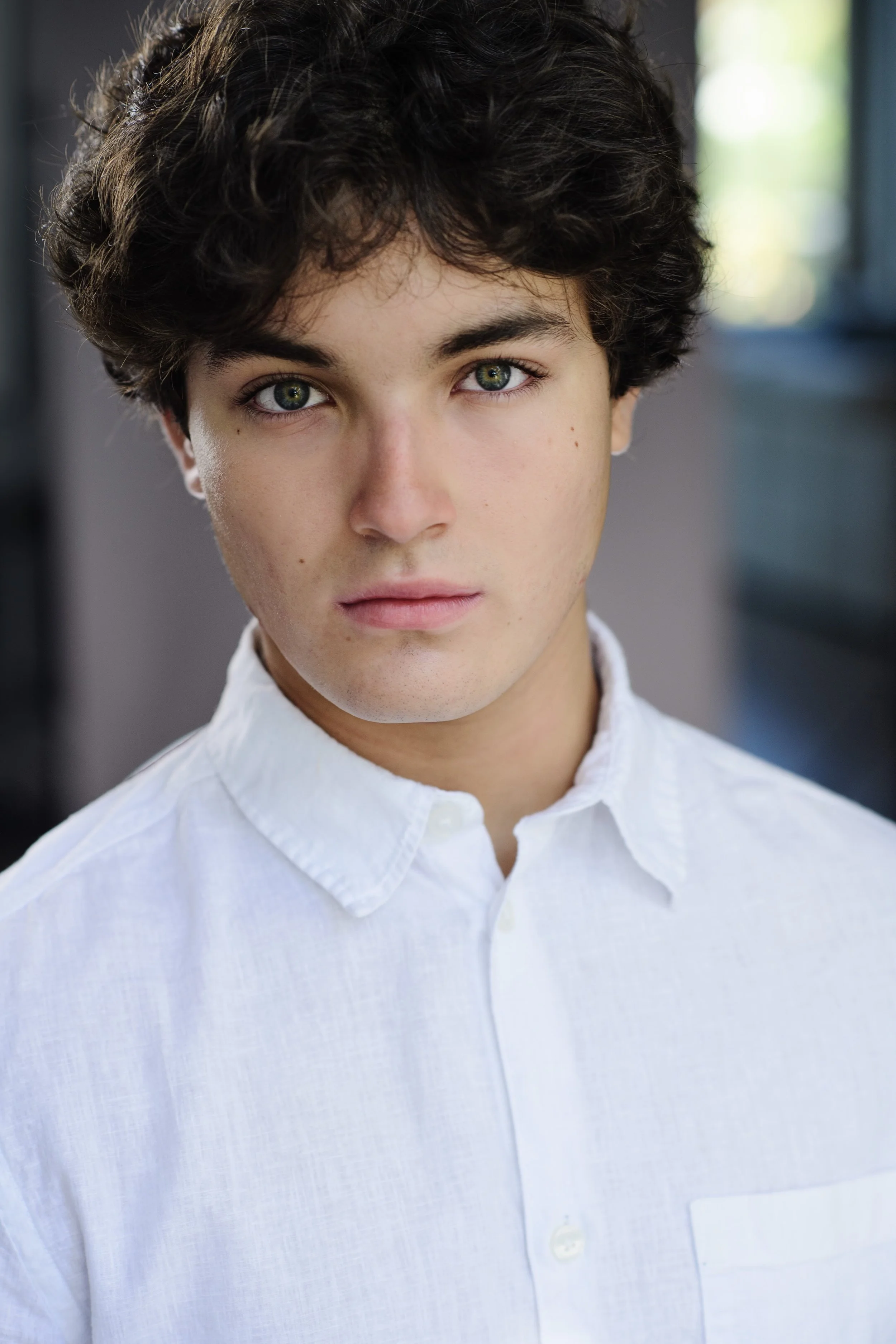 Close-up portrait of a young person with curly dark hair and green eyes, wearing a white collared shirt, standing indoors with blurred windows in the background.