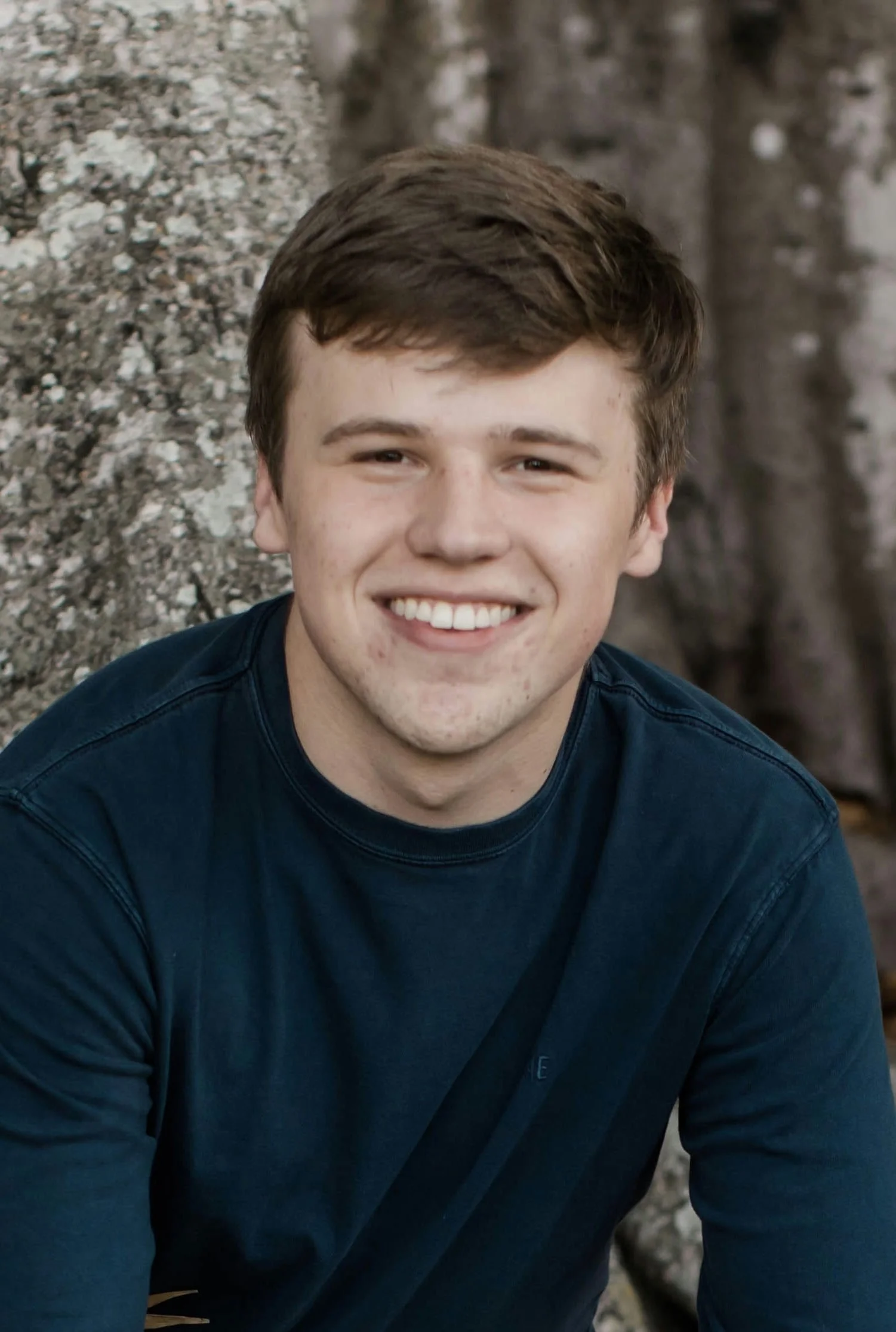 A woman with braided hair, wearing a blue sleeveless top, jewelry including earrings and necklaces, in an outdoor setting with blurred trees and grass in the background.