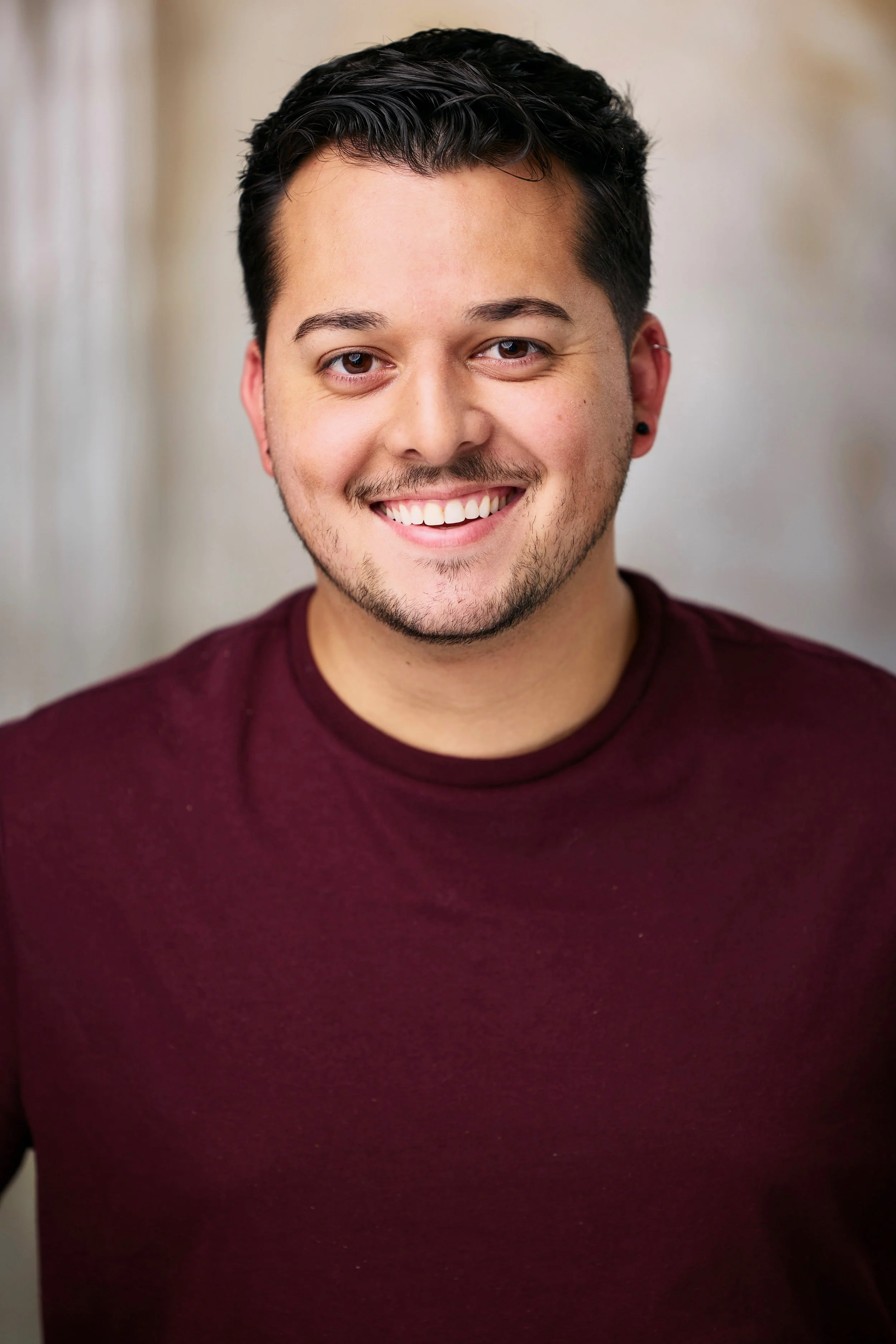 A young man with dark hair, light skin, and a beard, smiling and wearing a maroon shirt, in front of a soft, blurred background.