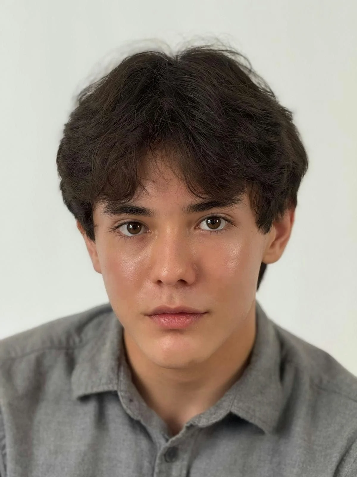Close-up portrait of a young man with dark wavy hair, light skin, and wearing a gray collared shirt, looking directly at the camera against a plain white background.