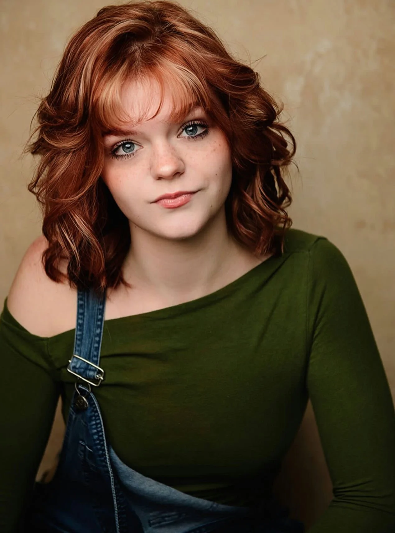 Close-up portrait of a young woman with long, wavy brown hair, green eyes, wearing a dark green top with a small bow at the neckline, smiling against a dark gray background.
