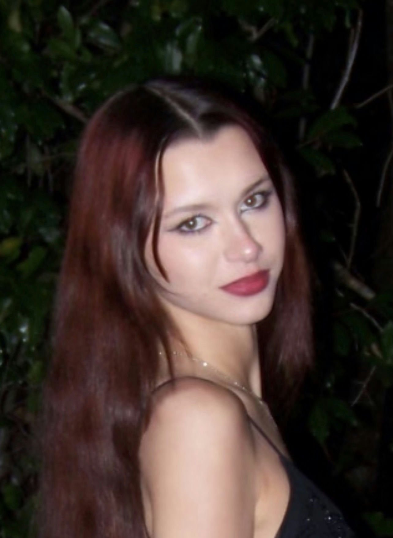 A young boy with tousled blond hair, wearing a maroon shirt, smiling at the camera against a plain light gray background.