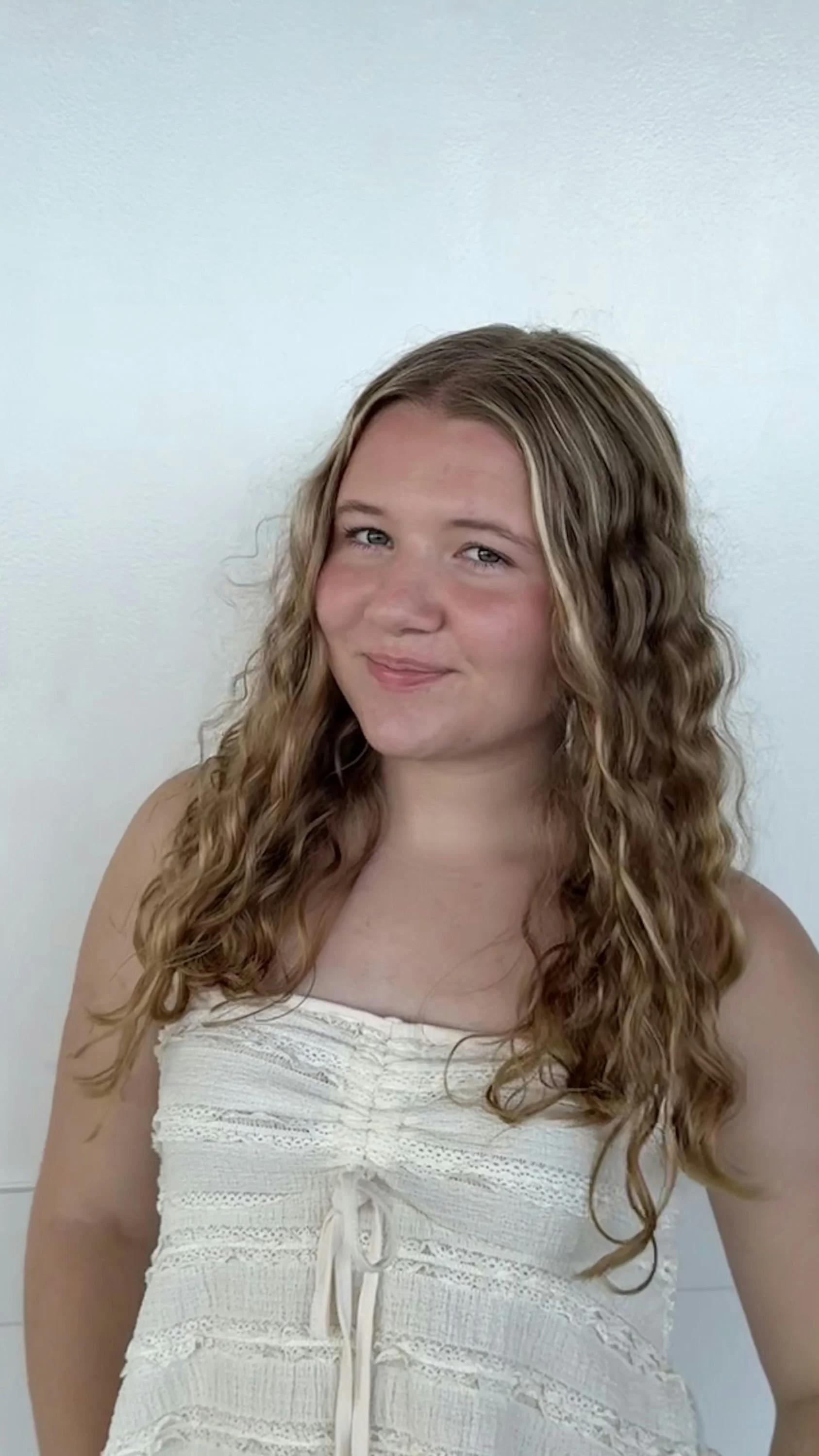 A young woman with long, wavy light brown hair and blue eyes, wearing a white sleeveless top, standing in front of a plain white wall, smiling softly at the camera.