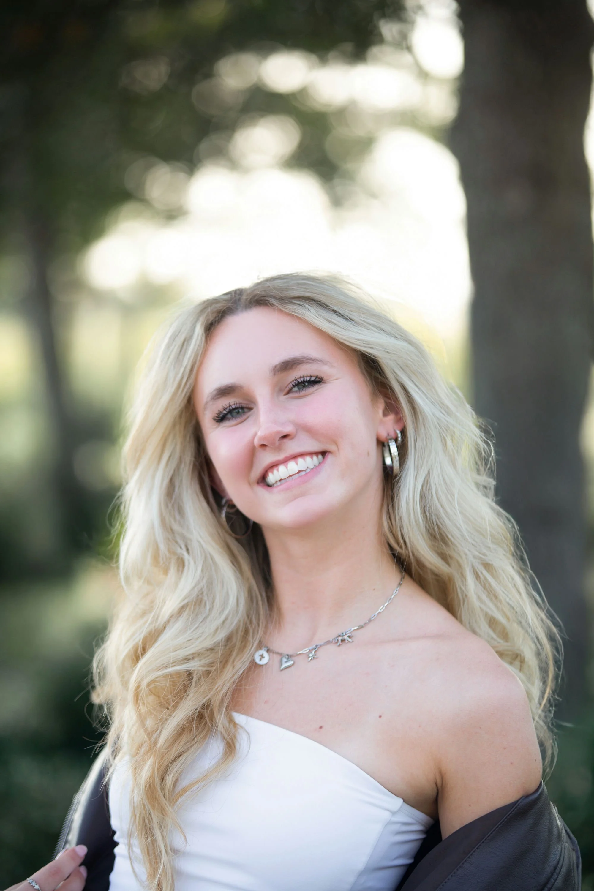 Portrait of a young woman with long blonde hair, smiling, outdoors with trees and sunlight in the background.